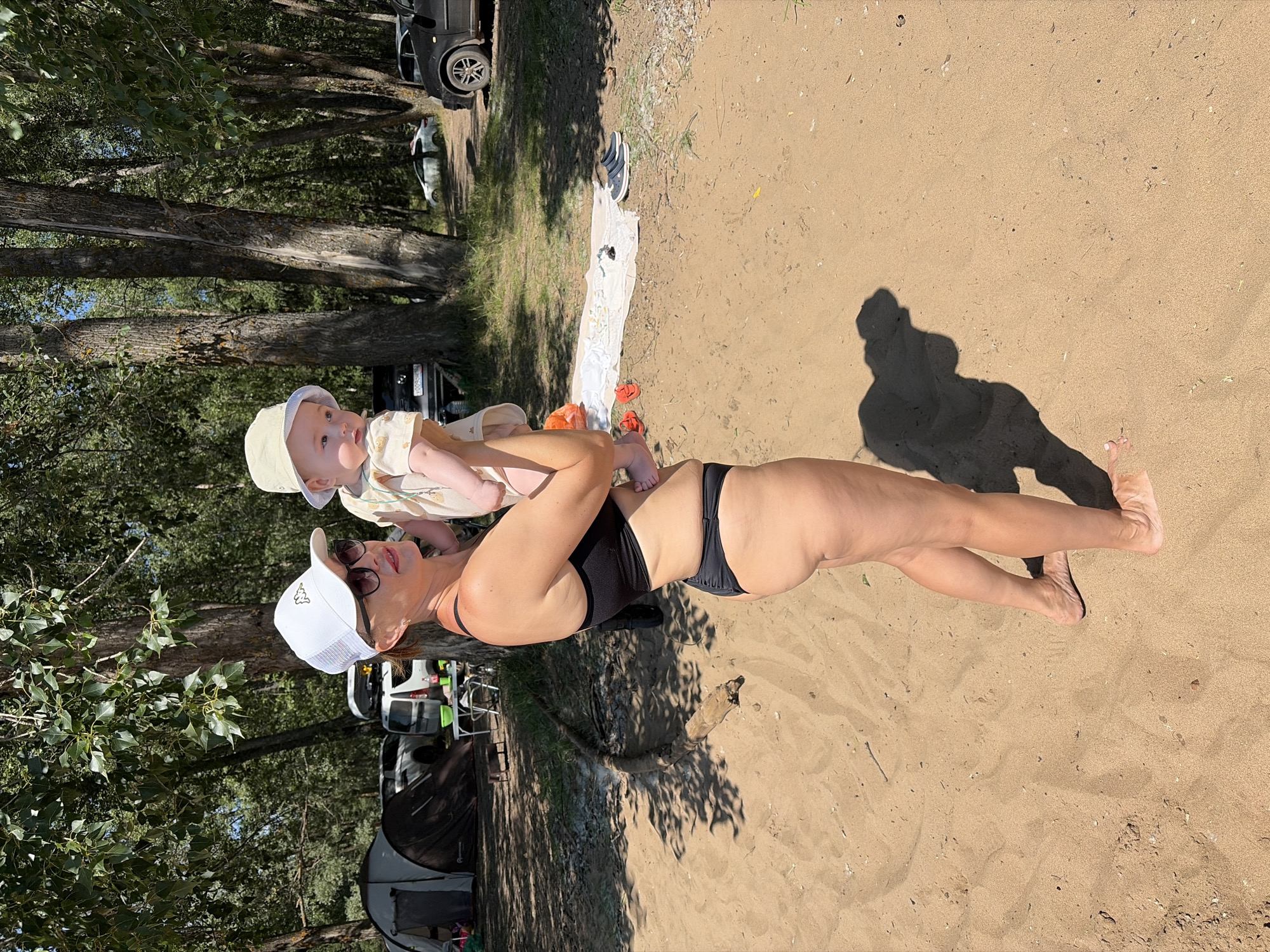 A woman in a black bikini and a white baseball cap holds a baby wearing a light-colored sun hat. They are standing on a sandy beach with trees and parked cars in the background, suggesting an outdoor recreational area, possibly near a body of water. The woman's bikini bottoms are black and thong-style, and she is wearing sunglasses. The baby is being held close to her chest. To the left, a dark tent is visible. In the middle ground, there's a table with items on it, and further back, several cars are parked. The ground is sandy, with some shadows from the trees cast across it. The scene appears to be in Noviy, Russia, based on the provided information.