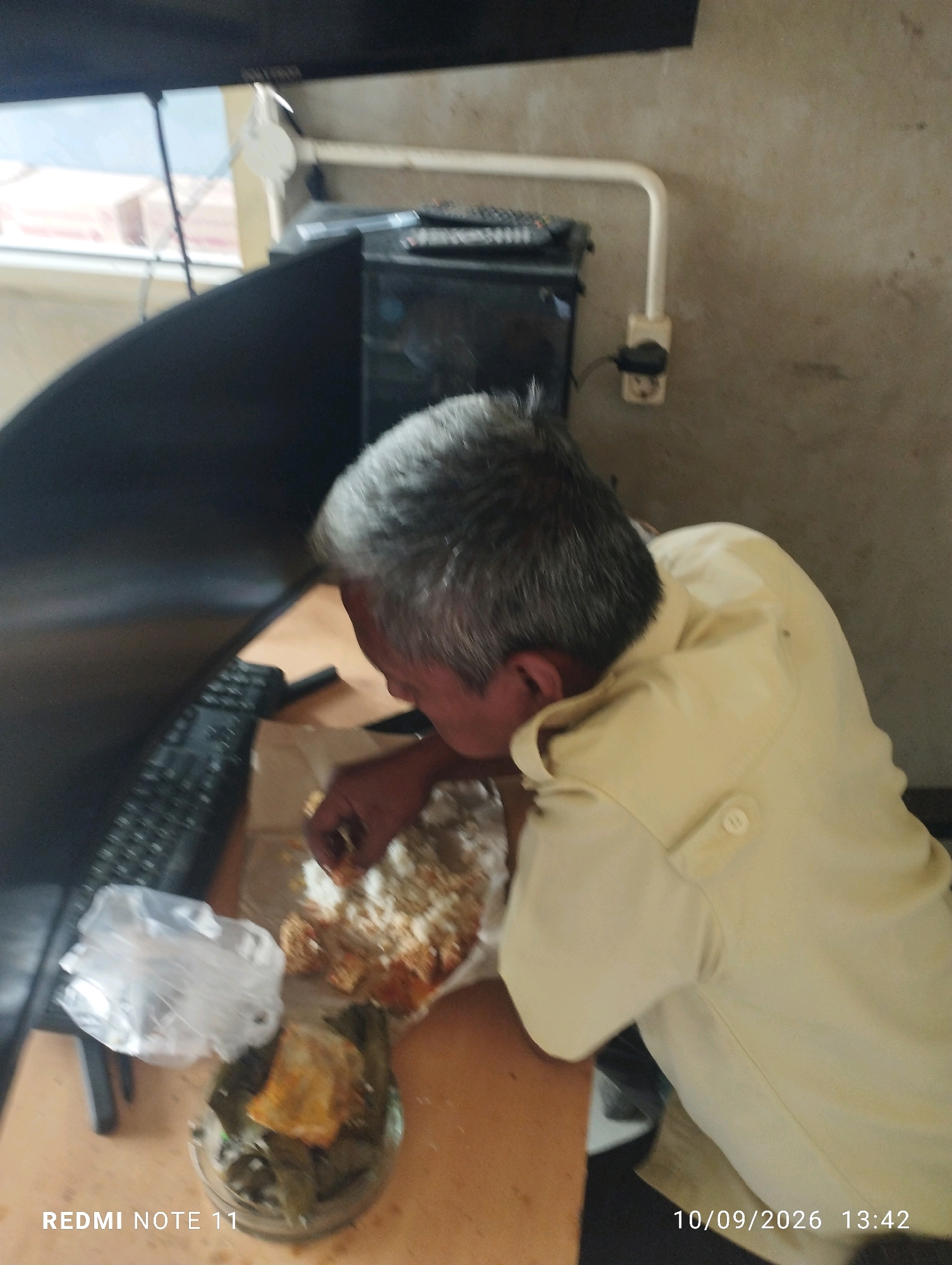The image shows a man with graying hair eating from a plate on a wooden desk. He is wearing a light-colored short-sleeved shirt. In front of him on the desk are several food items, including a plate of rice with some toppings, a clear plastic bag, and a clear bowl containing what appears to be food wrapped in leaves. Behind him, a black computer monitor is visible. A keyboard is also on the desk. In the background, a wall and a window can be seen. The timestamp "10/09/2026 13:42" is visible in the lower right corner, and "REDMI NOTE 11" is in the lower left corner. The scene appears to be indoors, possibly in an office or home setting. There is no indication of the location being in Bero, Indonesia.