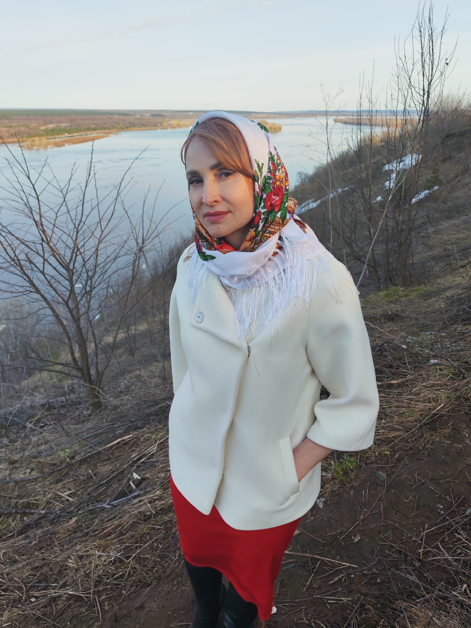 A woman stands on a hillside overlooking a river in Sarapul, Russia. She wears a white coat, a red skirt, and black boots. Her head is covered with a white and floral scarf with fringe. Her left hand is in her coat pocket. The hillside is covered with dry vegetation and bare trees. A river stretches out in the background, with land visible on the opposite bank. The sky is overcast.