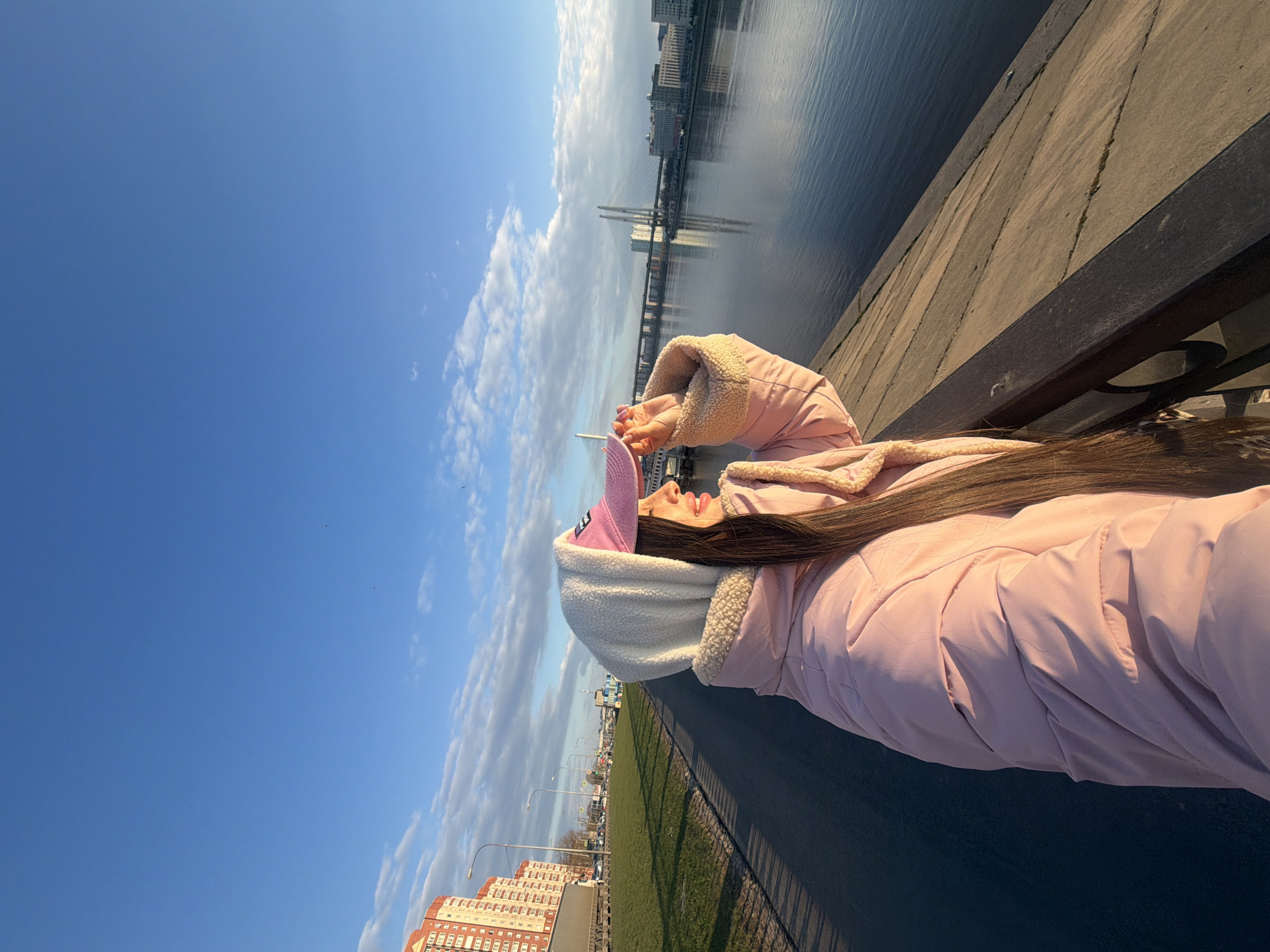 A woman wearing a pink jacket, a light-colored hooded sweatshirt, and a pink cap stands facing away from the camera, with a body of water and a cityscape in the background. Her long brown hair extends down her back. She is standing on a paved walkway with a low railing separating it from the water.
The cityscape includes several tall buildings and a cable-stayed bridge spanning the water.
The location is Saint Petersburg, Russia. The cityscape in the background includes the Lakhta Center and the Saint Petersburg Stadium. The bridge is the Cable-Stayed Bridge, and the body of water is either part of the Neva River or the Gulf of Finland. The buildings in the background are located along the Primorsky District.