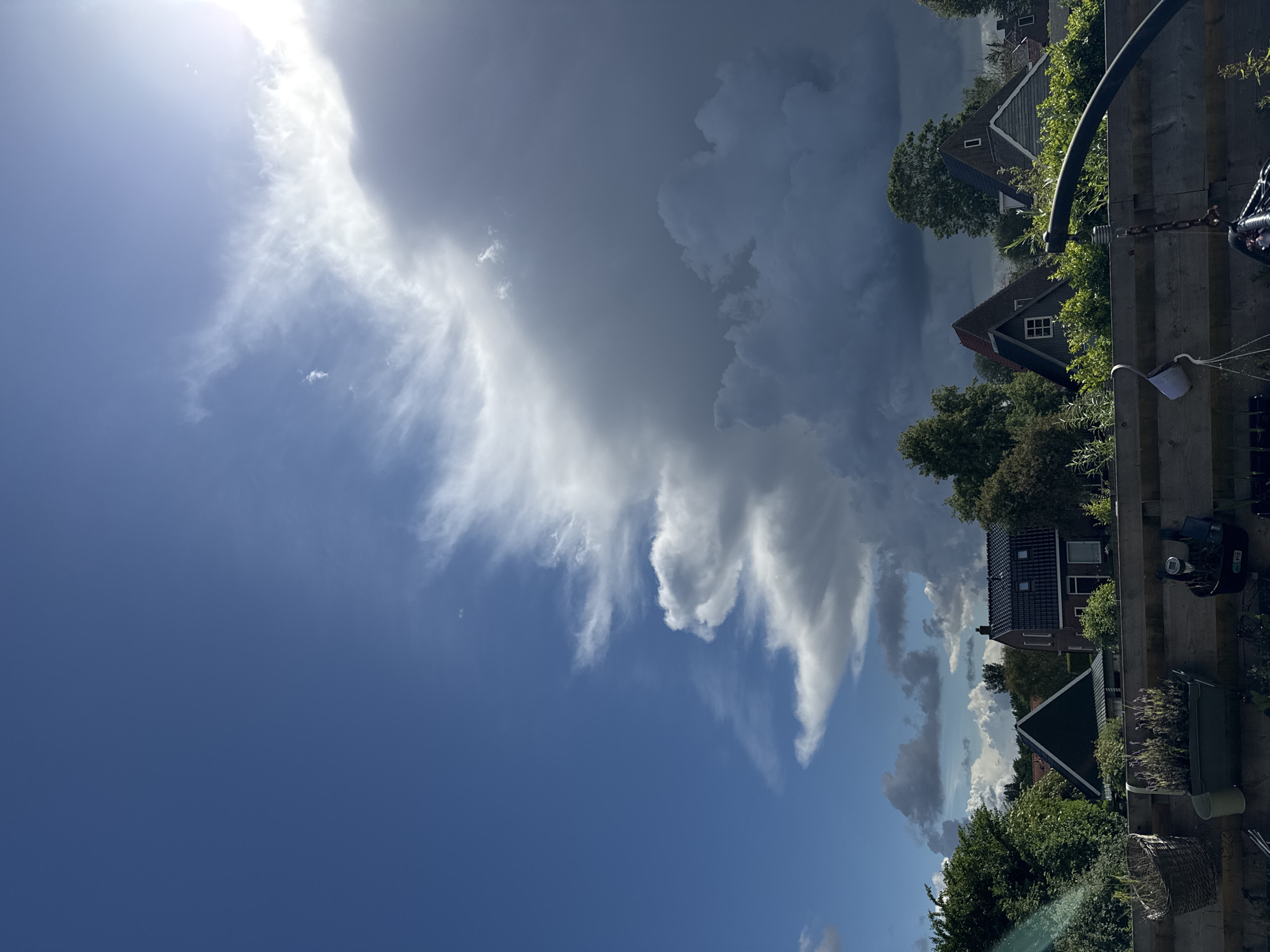 A scene looking up at a dramatic sky above a residential area. The sky is split between a bright blue expanse on the left, with wispy white clouds, and a large, dark, stormy cloud bank on the right, illuminated by the sun from the upper right corner. Below the sky, the tops of several houses with pitched roofs are visible, interspersed with lush green trees and bushes. In the foreground, a wooden fence runs horizontally across the bottom of the frame, with several potted plants and various objects hanging from it, including a basket, a watering can, and what appears to be part of a swing set. The lighting suggests it is daytime, with the sun casting some shadows.