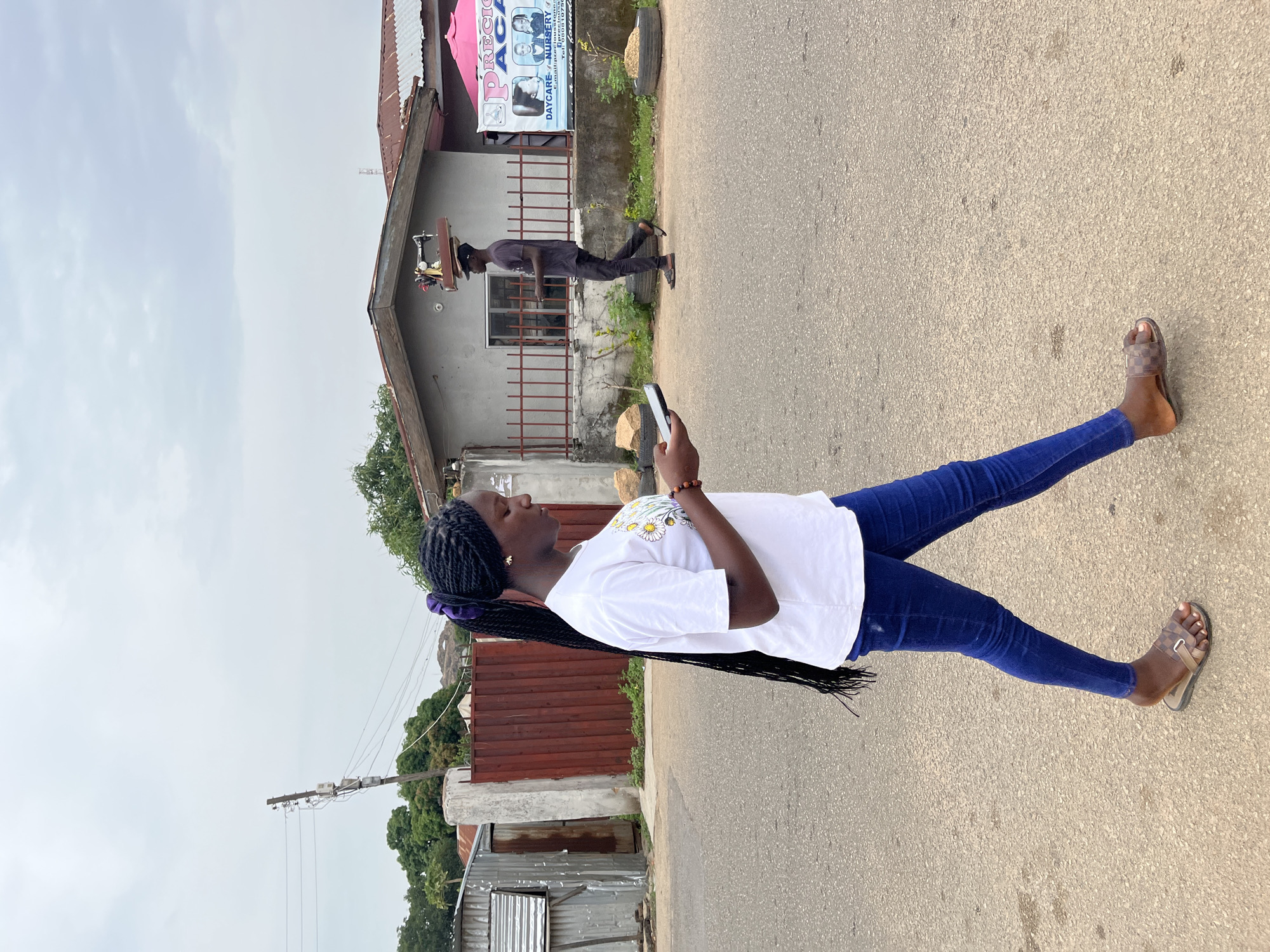 This outdoor street scene, likely in Jos, Nigeria, features a young woman in the foreground and a man in the background. The main subject, a woman with long black braids tied with a purple scrunchie, stands on an asphalt road, looking down at a smartphone she holds in both hands. She wears a white t-shirt with yellow and white floral patterns, blue jeans, and light brown patterned sandals.
In the mid-ground, a man walks away from the viewer, expertly balancing a table-mounted sewing machine on his head. He is dressed in dark attire and sandals. The background consists of several buildings, including a concrete structure with a brown metal fence. Above this building, a partially visible pink banner reads "PRECI...", "DAYCARE + NURSERY", and part of "a sure foundation", accompanied by images of children. Planters made from discarded tires are also visible.
The sky is overcast, suggesting daytime with diffuse lighting. The general setting includes residential or commercial buildings, corrugated metal structures, green foliage, and a utility pole with wires in the distance. The image portrays a candid moment of daily life in an urban or semi-urban environment.