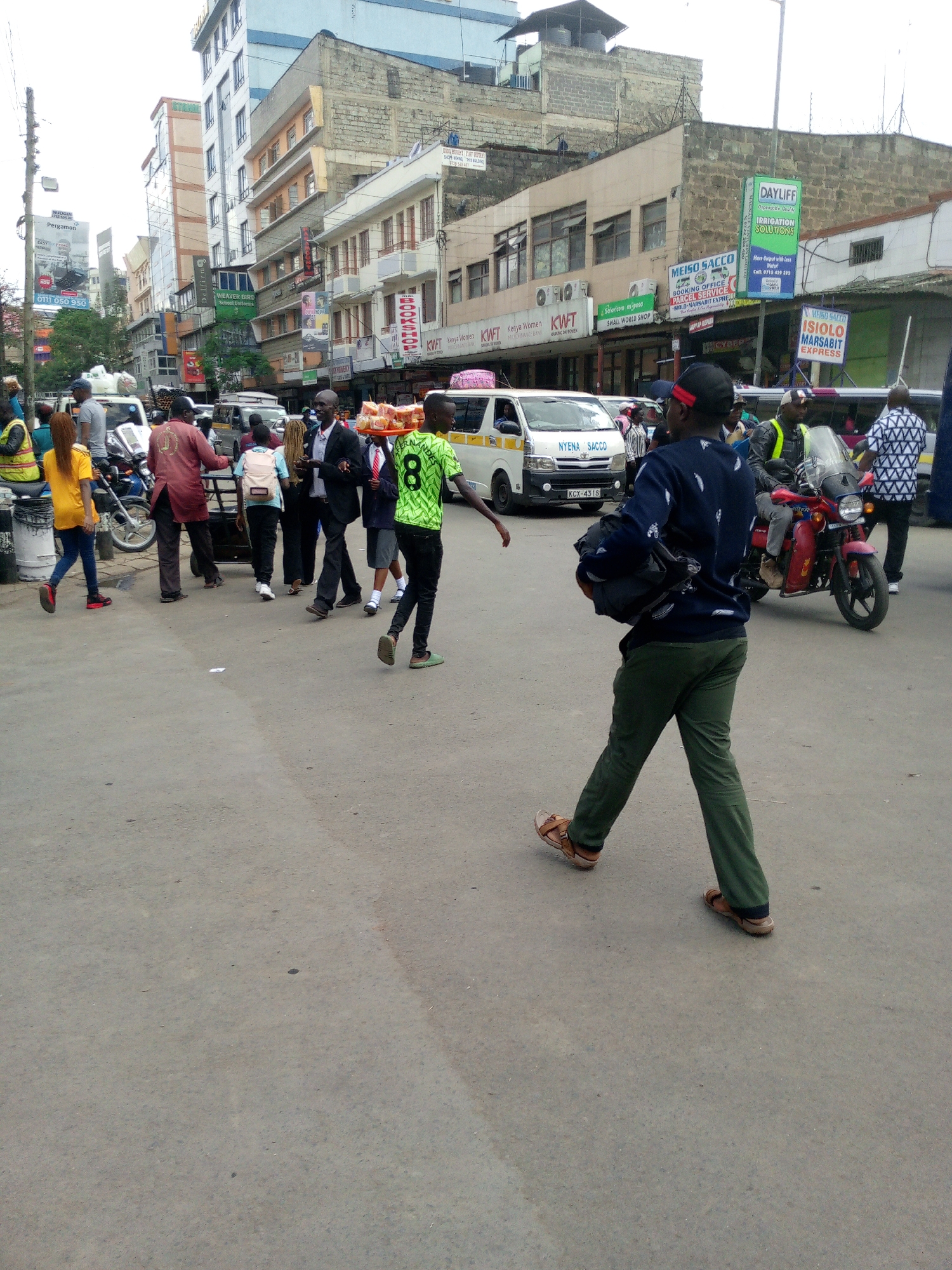 The image captures a bustling street scene in Nairobi, Kenya. In the foreground, a man wearing a dark blue sweater, green pants, and sandals walks away from the camera, carrying a dark-colored bag over his shoulder. In the middle ground, a group of people is crossing the street. One person, wearing a green shirt with the number 8 on the back, balances a tray of food on their head. Nearby, a man in a red jacket pushes a handcart. A yellow-shirted woman walks towards the left of the frame. A motorcycle, red in color, is parked with its driver and a passenger. A white van labeled "NYENA SACCO" is visible in the background. Several buildings are in the backdrop with commercial signs; the signs indicate businesses and services in the area. Signs such as "DAYLIFF IRRIGATION SOLUTIONS", "MEISO SACCO", and “ISIOLO MARSABIT EXPRESS" are clearly visible. Another sign indicates "KWFT Kenya Women". The road surface appears to be asphalt.