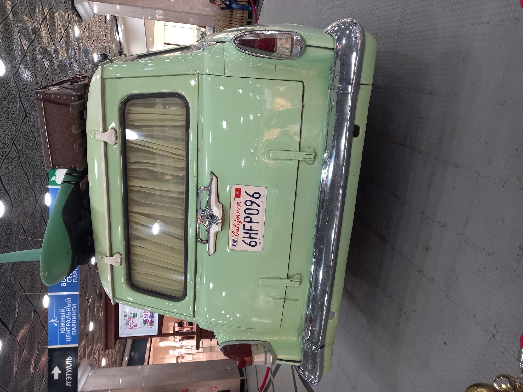 This image shows the rear of a light green vintage car parked indoors. The car has a California license plate that reads "6HFP096". On the roof of the car, there is luggage and a green object that resembles a surfboard or a plane wing. The car is parked on a gray, tiled floor. In the background, there are signs with Russian text, including one that reads "ЮЖНЫЙ, ЦЕНТРАЛЬНЫЙ ПАРКИНГ" which translates to "SOUTH, CENTRAL PARKING". These signs suggest the location is a parking area within a shopping mall or similar complex in Yekaterinburg, Russia. The ceiling of the space has a textured, patterned design. A person is visible sitting on a bench in the far right background.