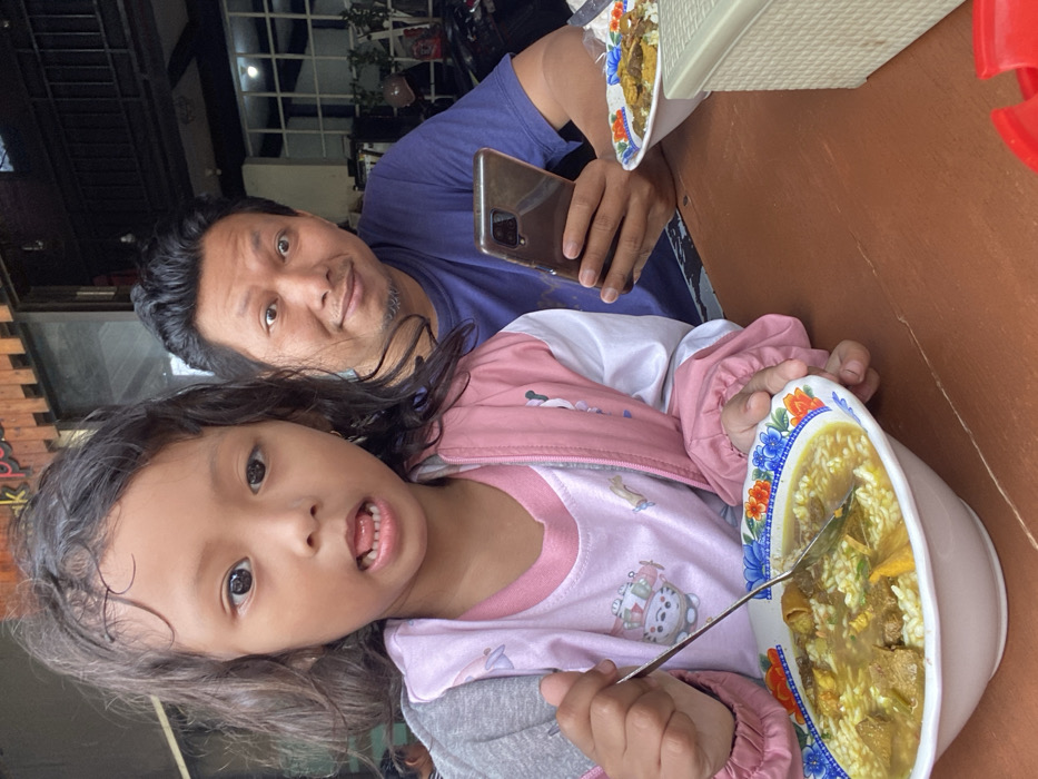 A man with short black hair and a blue shirt looks at a phone while a young girl eats soup. She wears a pink and gray jacket. The table is brown, and another plate of food is visible in the upper right. The image appears to be taken from above and rotated, as the man's head is tilted.