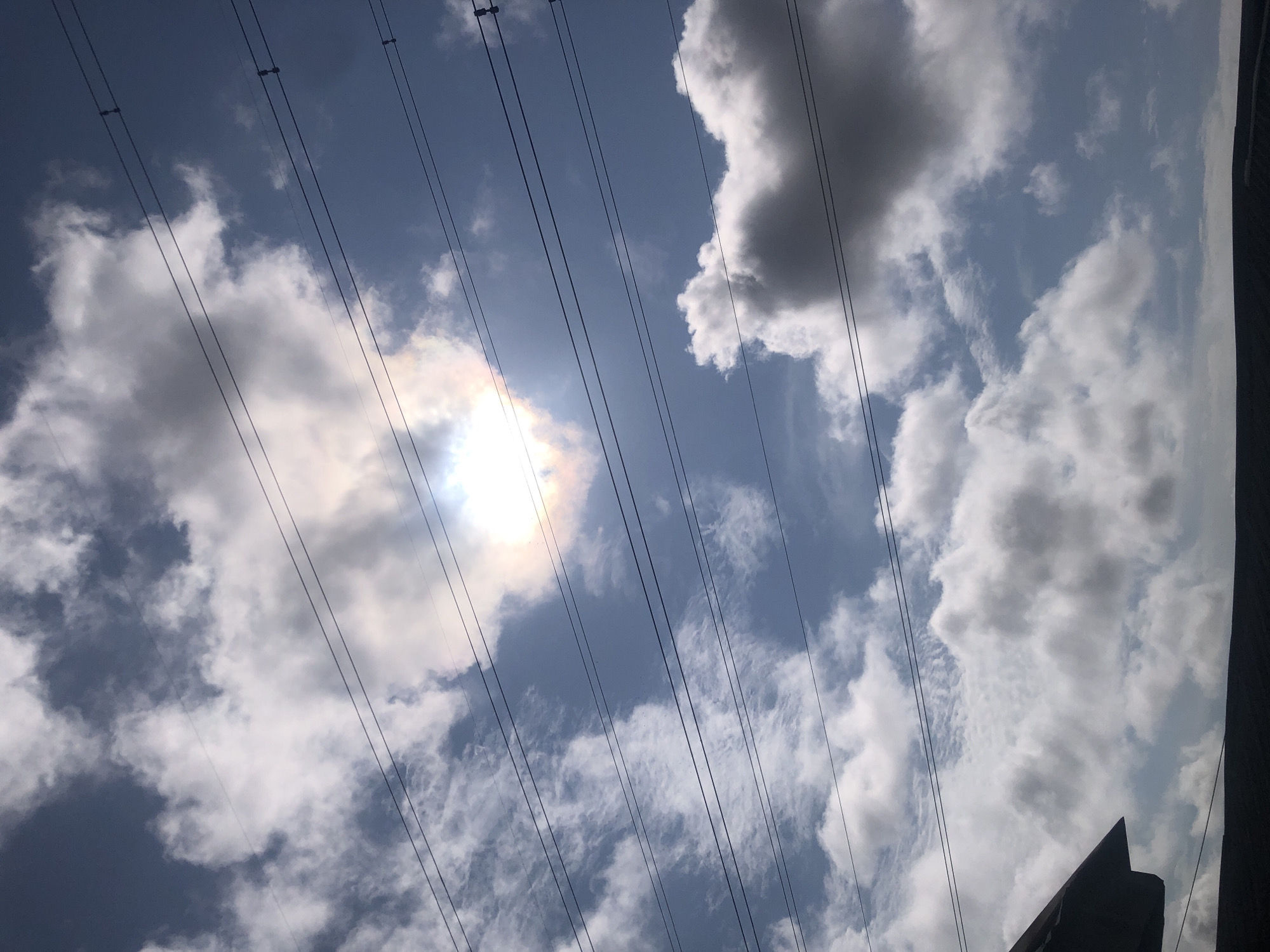 An outdoor, low-angle, eye-level shot captures a cloudy sky with power lines in Ajah, Nigeria. The sun is partially visible, creating a bright halo effect through a gap in the clouds. The sky is a mix of bright blue and white clouds, with some darker gray clouds present. Several parallel power lines stretch across the frame from left to right. The top left corner of the image shows the dark roofline of a building. The bottom of the image features a dark, undefined horizontal structure, possibly another building or a fence. The scene is devoid of any people or animals.