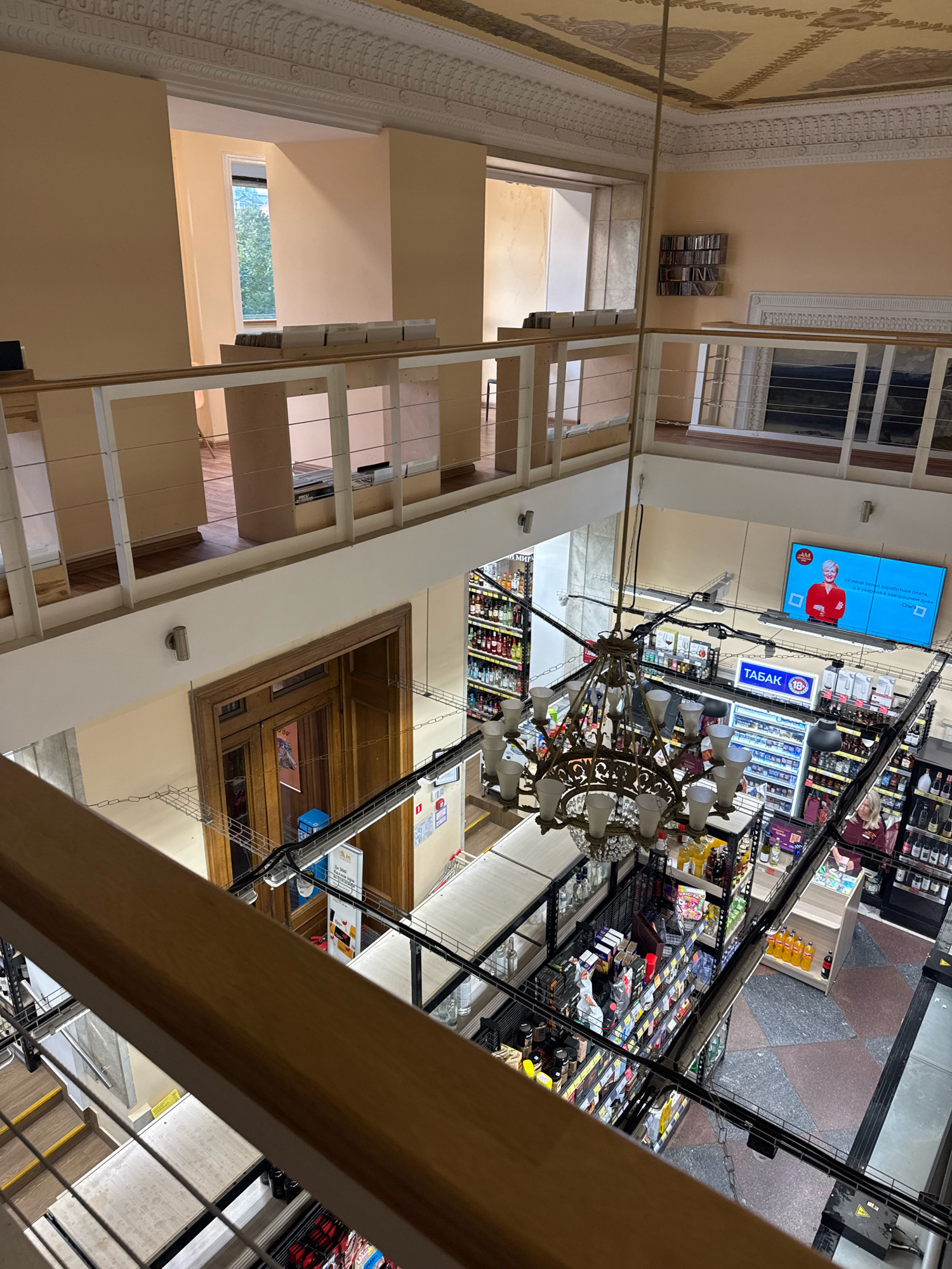 This is an overhead, wide-angle photograph taken from a higher level, looking down into a multi-story interior space. The architecture features ornate decorative molding on the ceiling and walls, suggesting a historic building. A prominent element is a large, elaborate chandelier suspended from the ceiling, illuminating the space.
The lower level appears to be a retail establishment, possibly a supermarket or convenience store, with aisles of shelves stocked with various products, including beverages and packaged goods. A digital display screen is mounted on a wall, showing a woman in a red shirt and text in Russian. There are also signs in Russian, one of which reads "ТАБАК" (tobacco). A cashier, a woman with blonde hair wearing a maroon uniform, is visible behind a counter on the right side of the image.
The upper level has a balcony or walkway with a wooden railing, overlooking the retail area. A tall wooden door with glass panels is visible on the left side of the lower level. The overall impression is a blend of historical architecture and modern retail. The presence of Russian text and the style of the retail space suggest this location is in Moscow, Russia.
