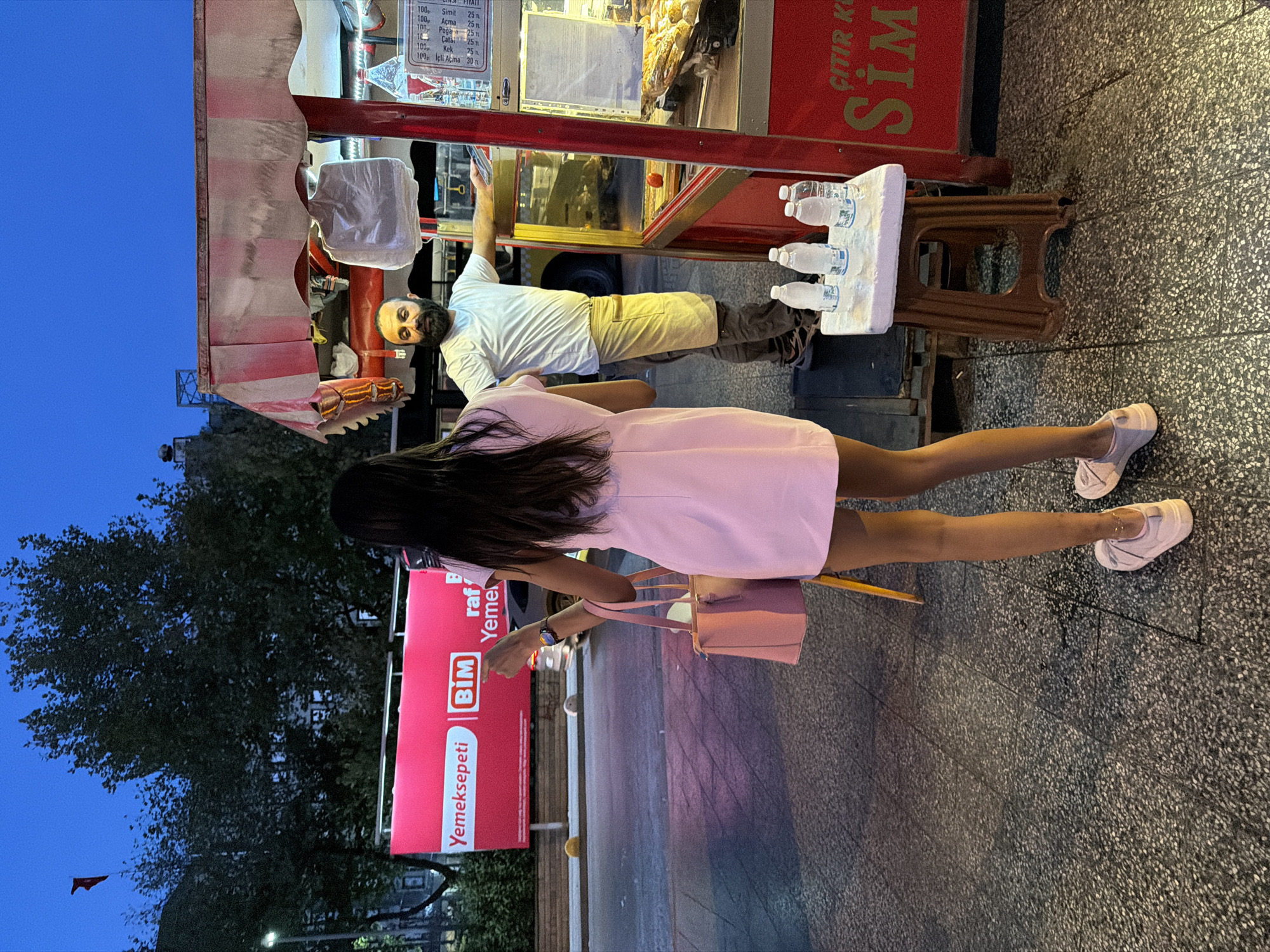 A man with a beard and a white t-shirt stands behind a food stall. He is facing a young woman in a pink dress, who is facing away from the camera and looking towards the stall. The food stall has a red and white striped awning and displays food items behind glass. A small stool with four bottles of water sits in front of the stall. In the background, a large red billboard with "Yemeksepeti" and "BIM" logos is visible. The scene appears to be a street in Kadıköy, Turkey, at dusk or early evening, as indicated by the dim lighting and the blue sky.
