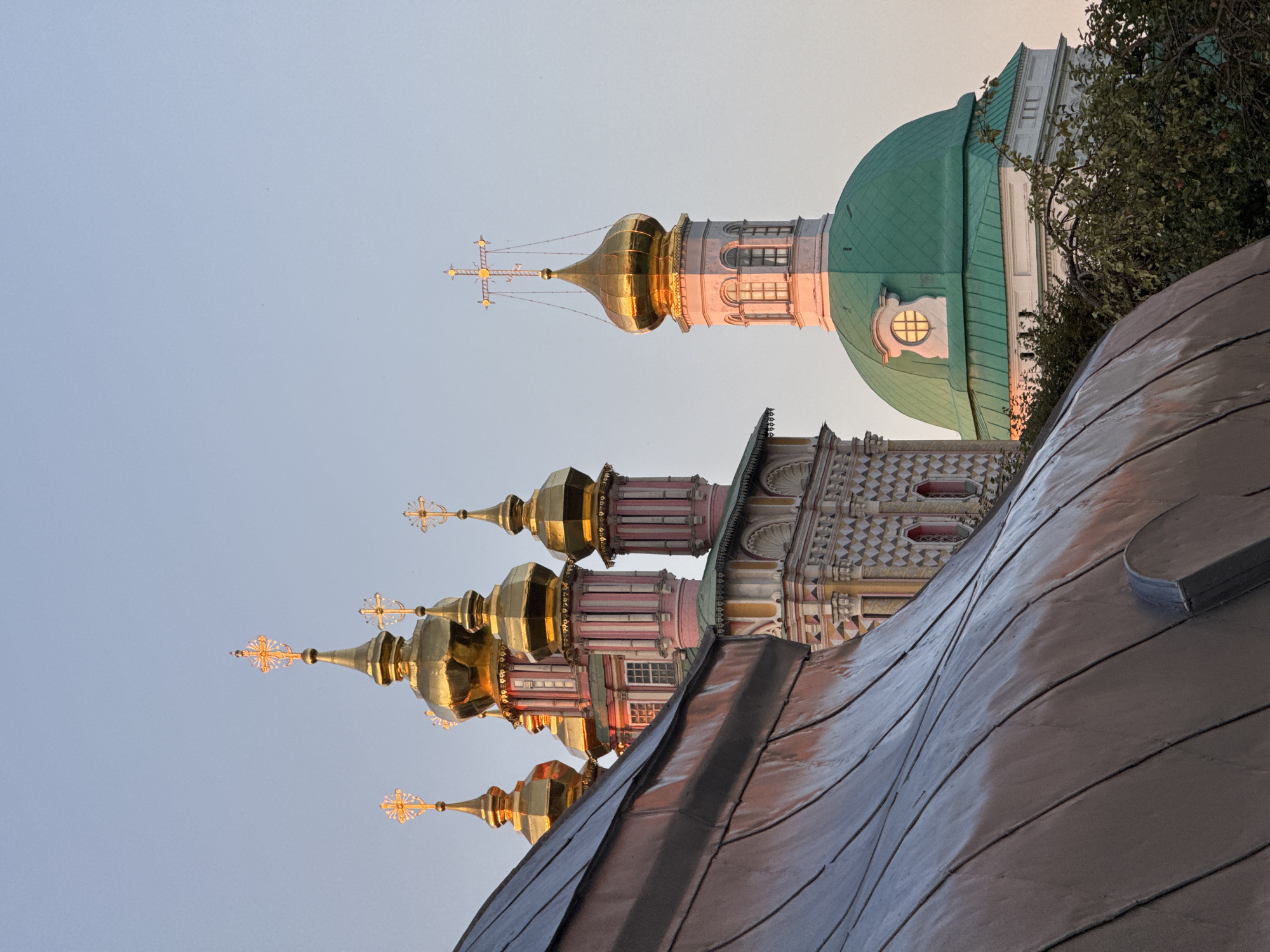 The image shows the Trinity Lavra of Saint Sergius in Sergiev Posad, Russia. In the foreground, a curved, weathered metal roof dominates the left side of the frame. Behind it, a cluster of ornate church domes and steeples rises against a clear, pale sky. The domes are predominantly gold, topped with intricate crosses. The steeples are painted in pastel pink and white with decorative elements. To the right, a larger, green dome with a circular window is visible, also adorned with a cross at its peak. The lighting suggests it is either sunrise or sunset, casting a warm glow on the golden domes. Trees with green foliage are visible in the lower right corner.