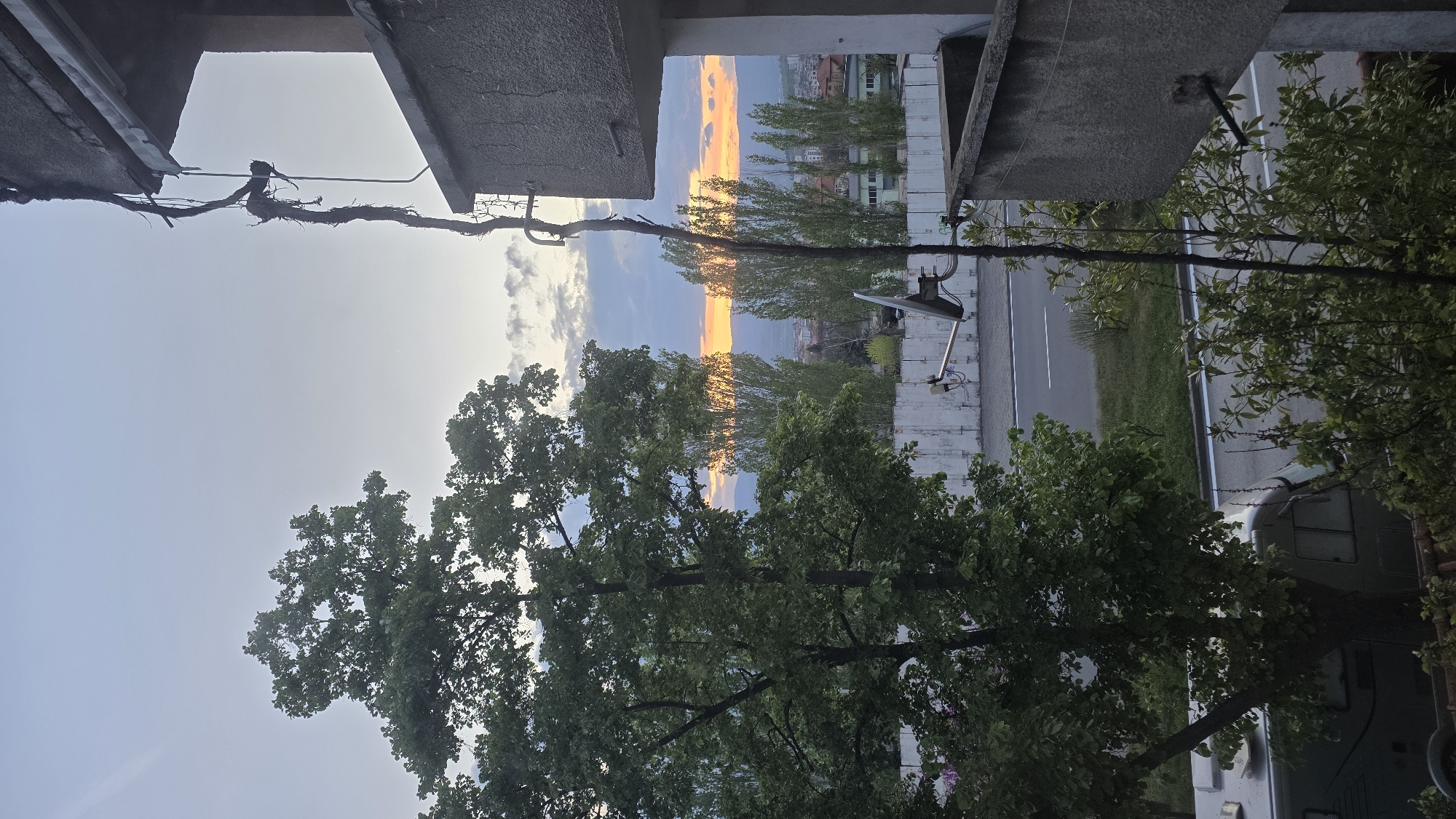 The image captures an urban vista at sunset, viewed from a high vantage point, likely a balcony or window in Blagoevgrad, Bulgaria.
In the immediate foreground, concrete architectural elements, possibly parts of a building's structure, frame the top and bottom of the scene. A dark, thick, and tangled cable or vine stretches across the upper part of the image, connecting these structures. Lush green trees dominate the lower foreground and extend up both sides of the frame, their foliage partially obscuring other elements.
The midground features a paved road with white lane markings running horizontally. Adjacent to the road are grassy verges and more trees. On the right side of the road, a light-colored vehicle, possibly a truck or a large van, is partially visible. A white satellite dish or security camera array is mounted on the concrete structure to the right.
The background is dominated by a dramatic sunset. A brilliant horizontal band of intense orange and yellow light illuminates the distant sky, indicating late afternoon or early evening. Above and below this vibrant strip, the sky transitions to shades of blue and grey, with some scattered clouds. In the far distance, hills or mountains are silhouetted against the colorful sky, with glimpses of a town or city nestled amongst them.
No specific activity is taking place; the image presents a serene urban landscape bathed in the warm glow of dusk. There is no visible text.