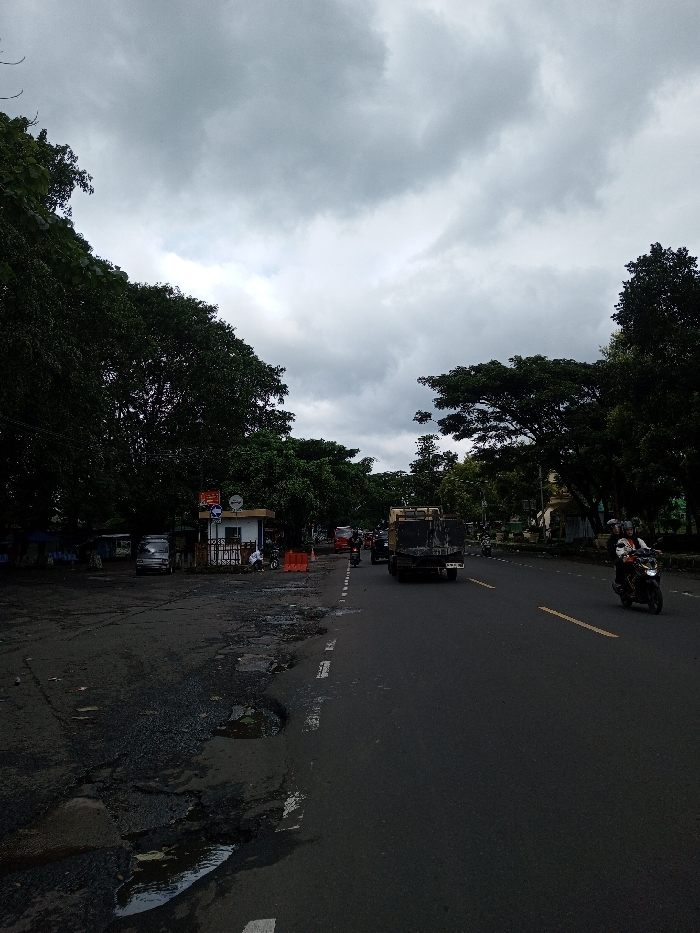 The image depicts an outdoor street scene under a heavily overcast sky, suggesting a cloudy day, possibly before or after rain, given the puddles on the ground.
The main subject is a two-lane asphalt road, which stretches from the foreground into the distance. The road appears dark grey and has clear white dashed lines marking the center or edge of the lanes, along with some yellow lines visible further down. To the left of the main road, there's an unpaved, dirt and gravel shoulder that is in poor condition, marked by numerous large potholes, some of which are filled with water. This area seems to be a common space for parking and potentially small makeshift stalls.
On the left side of the road, a line of dense, dark green trees forms a canopy, creating a shadowed area beneath them. Beneath these trees, in the mid-ground, there's a small, light-colored building, possibly a guard post or a small kiosk. On top of this building, a blue sign is visible with white text, partially readable as "Selamat Datang" (which translates to "Welcome" in Indonesian/Malay). Below this text, there's a white circular logo or icon. Several vehicles are parked near this building, including a white van. There are also what appear to be makeshift structures or stalls with blue tarps further back.
On the right side of the road, another line of large, dark green trees also lines the street, obscuring distant buildings or structures. A white curb or barrier runs along the edge of the paved road.
Vehicles are visible on the road. In the middle of the road, a dark-colored dump truck or utility truck is traveling away from the viewer. Behind it and further down the road, other vehicles, including several motorcycles and possibly cars, can be seen. Closer to the foreground on the right side of the road, a motorcycle with two occupants (a driver and a passenger, both wearing helmets) is riding towards the viewer, or slightly angled away. Other people are visible near the small building on the left.
The sky occupies the upper portion of the image and is filled with various shades of grey clouds, with some lighter patches indicating where the cloud cover might be thinner. The overall lighting is diffused, consistent with an overcast day.