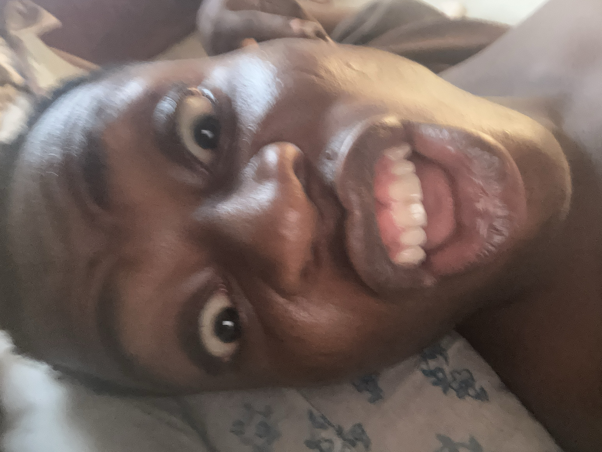 A close-up of a Black person's face, lying on a bed, looking up at the camera with their mouth open, revealing their teeth. The background consists of bedding with a blue and white pattern. The location is unknown.