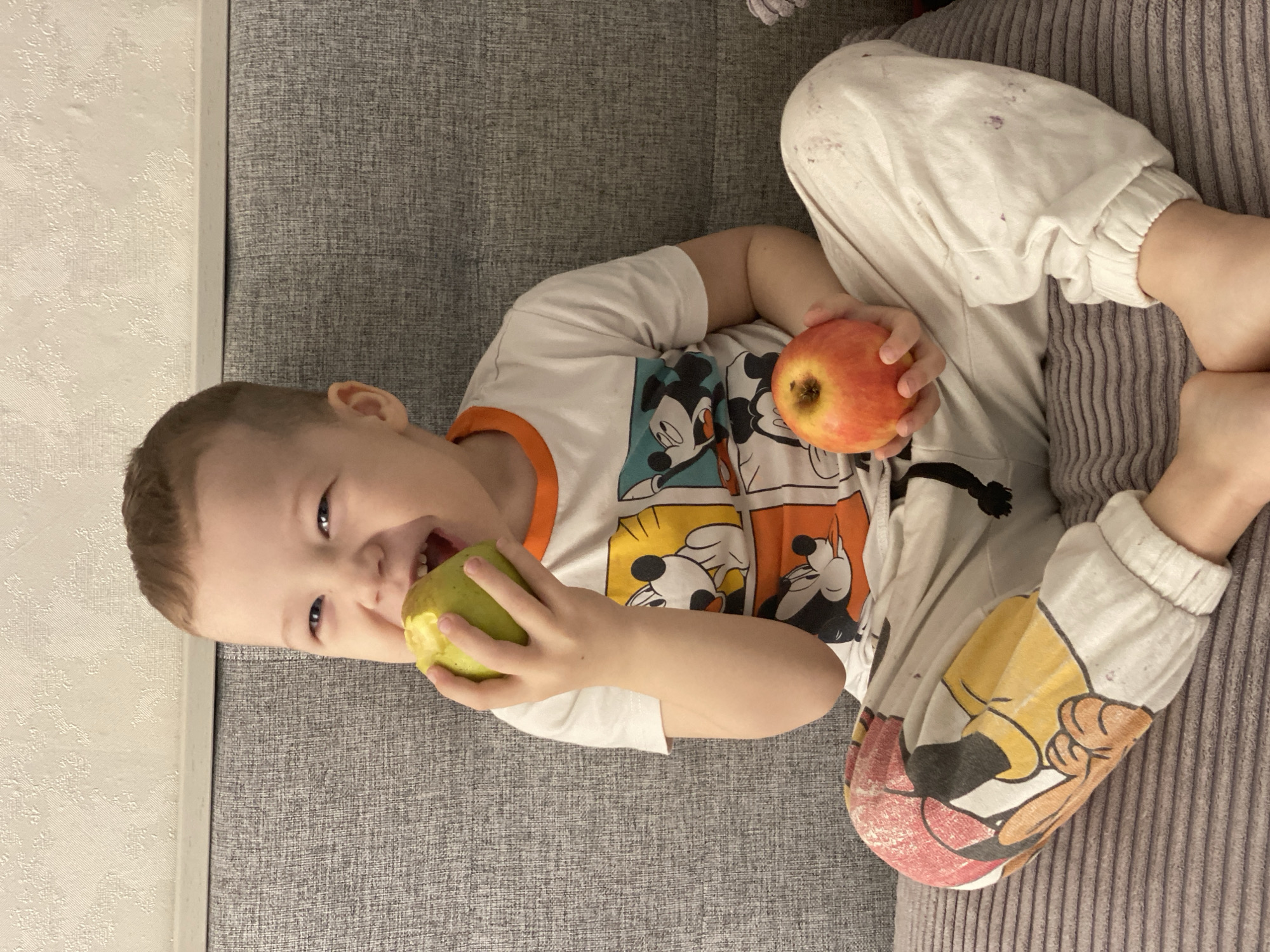 A young boy is lying on a gray textured sofa, possibly indoors. He is wearing a white t-shirt with a cartoon character print and matching white pants. He is holding a green apple in his left hand, taking a bite, and a red apple in his right hand. His expression is one of enjoyment. The location, Sochi, Russia, is not discernible from the image.