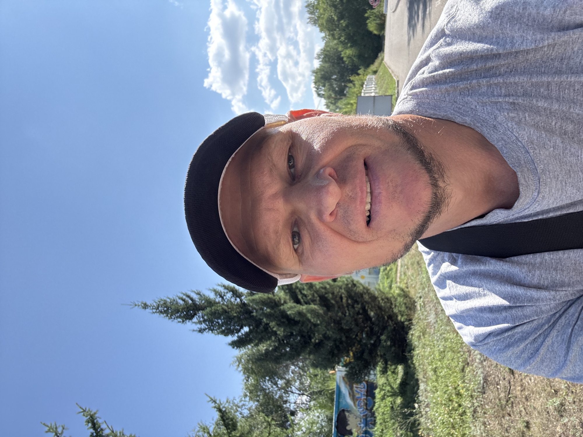 The image is a close-up selfie taken outdoors on a sunny day. The subject is a man wearing a grey t-shirt and a black baseball cap, with a black strap visible across his shoulder. He is smiling and looking directly at the camera. In the background, there are green trees, a bright blue sky with scattered clouds, and a paved road or path. To the left of the man, there is a section of a mural or graffiti on a wall, depicting animated figures. The man's attire and the overall setting suggest he might be in a park or an outdoor recreational area. It is not possible to ascertain the specific location in the city of Oskemen, Kazakhstan from the image alone.