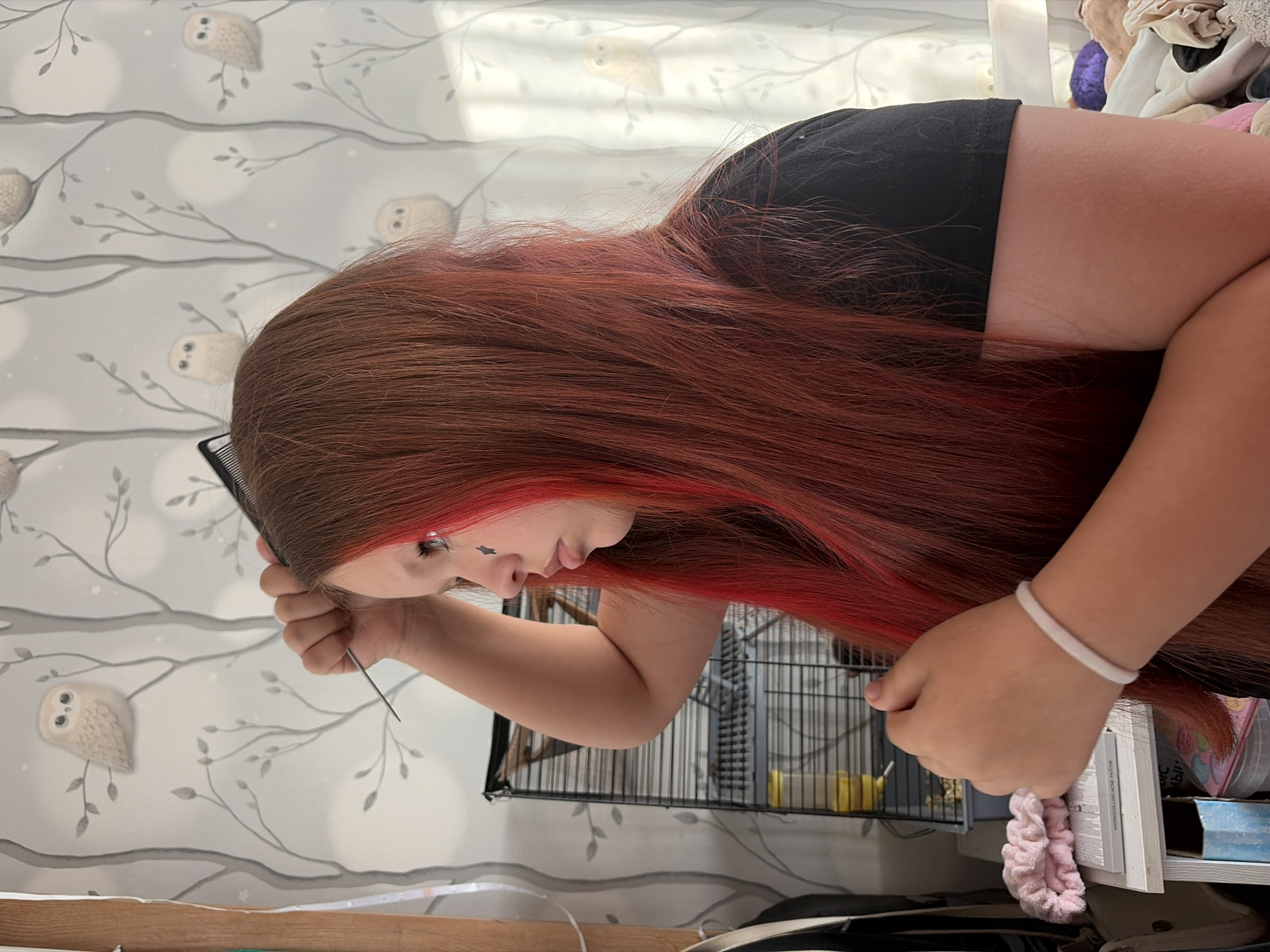 A fair-skinned individual with long, auburn hair is captured in a medium shot. The individual is wearing a black top and has a white bracelet on their left wrist. They are holding a comb in their right hand and appear to be styling or grooming their hair.
In the background, there is a wall with a pattern of stylized owls and branches. A wire cage with various objects inside, including what appears to be food and toys, is also visible. Additionally, there are some shelves with miscellaneous items, including a pink scrunchie and various containers. The location cannot be verified as being in Chaikovsky, Russia.