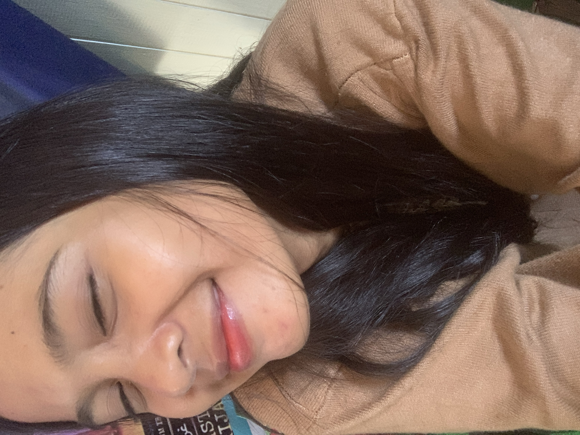 The image is a close-up, selfie-style photograph of a young woman. She has her eyes closed and is smiling. Her lips are tinted with a reddish-pink color, and she has long, dark hair that is partially obscuring her face. She is wearing a light brown, knitted sweater. The background is blurred and consists of vertical wooden planks and a dark blue object. There is also some text visible on the left side of the image, which appears to be from a poster or book. The lighting suggests an indoor setting. No specific location in Sungai Meriam, Indonesia, can be determined from the image.