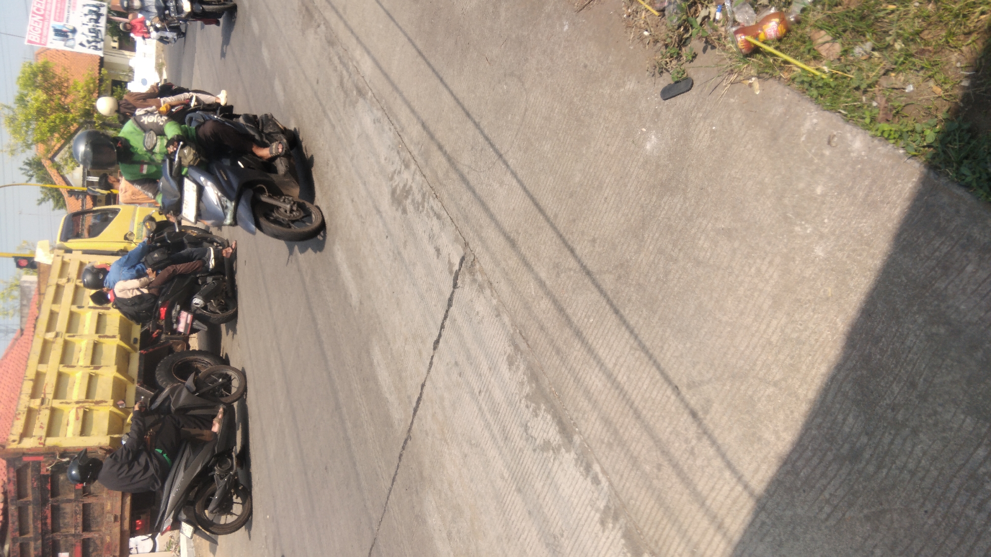 The image shows a street scene in Batang, Indonesia, with several motorcycles and a truck. In the foreground, a person wearing a green jacket and a black helmet is riding a dark-colored motorcycle. Behind them, another person in a blue shirt and a light-colored jacket is riding a motorcycle, carrying a passenger wearing a light-colored shirt and a black helmet. To the left, a person in a black shirt and helmet is riding a dark motorcycle. A large yellow truck is parked or moving in the background, next to a building with a red roof. Several other motorcycles are visible further down the street. The road is made of concrete with visible cracks and lines. Sunlight casts shadows on the road, indicating it is daytime. The area appears to be an urban environment.