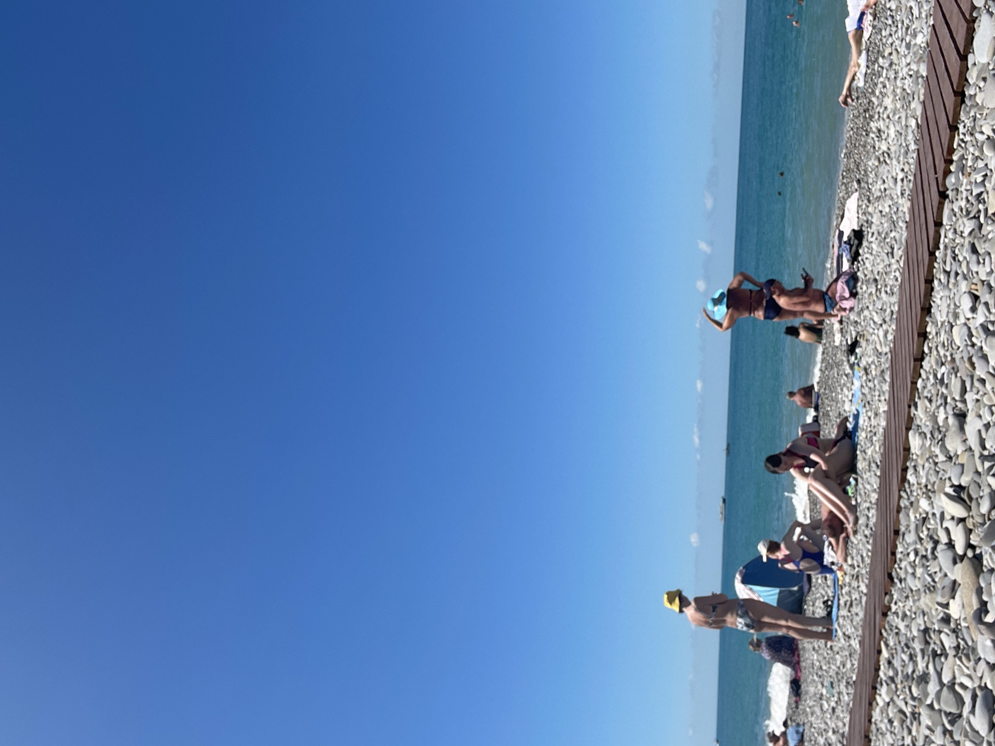 The image depicts a sunny day at a pebble beach. The sky is clear and blue. Several people are present, engaged in various beach activities. In the foreground, a wooden walkway runs horizontally across the pebble-covered shore.
To the left, a woman wearing a yellow hat and a patterned bikini stands on the beach. Nearby, a blue beach umbrella is set up, and other people are sitting on towels.
In the middle ground, a woman in a blue hat and bikini stands with her hand on her hip, looking out at the water. Beside her, a man in swimming trunks kneels, looking at his phone. A woman in a pink and black bikini sits on a towel, looking down.
Further out, the turquoise water of the sea extends to the horizon, where several boats are visible. There are also people in the water and further down the beach. The overall scene is one of a relaxed summer day at the beach.