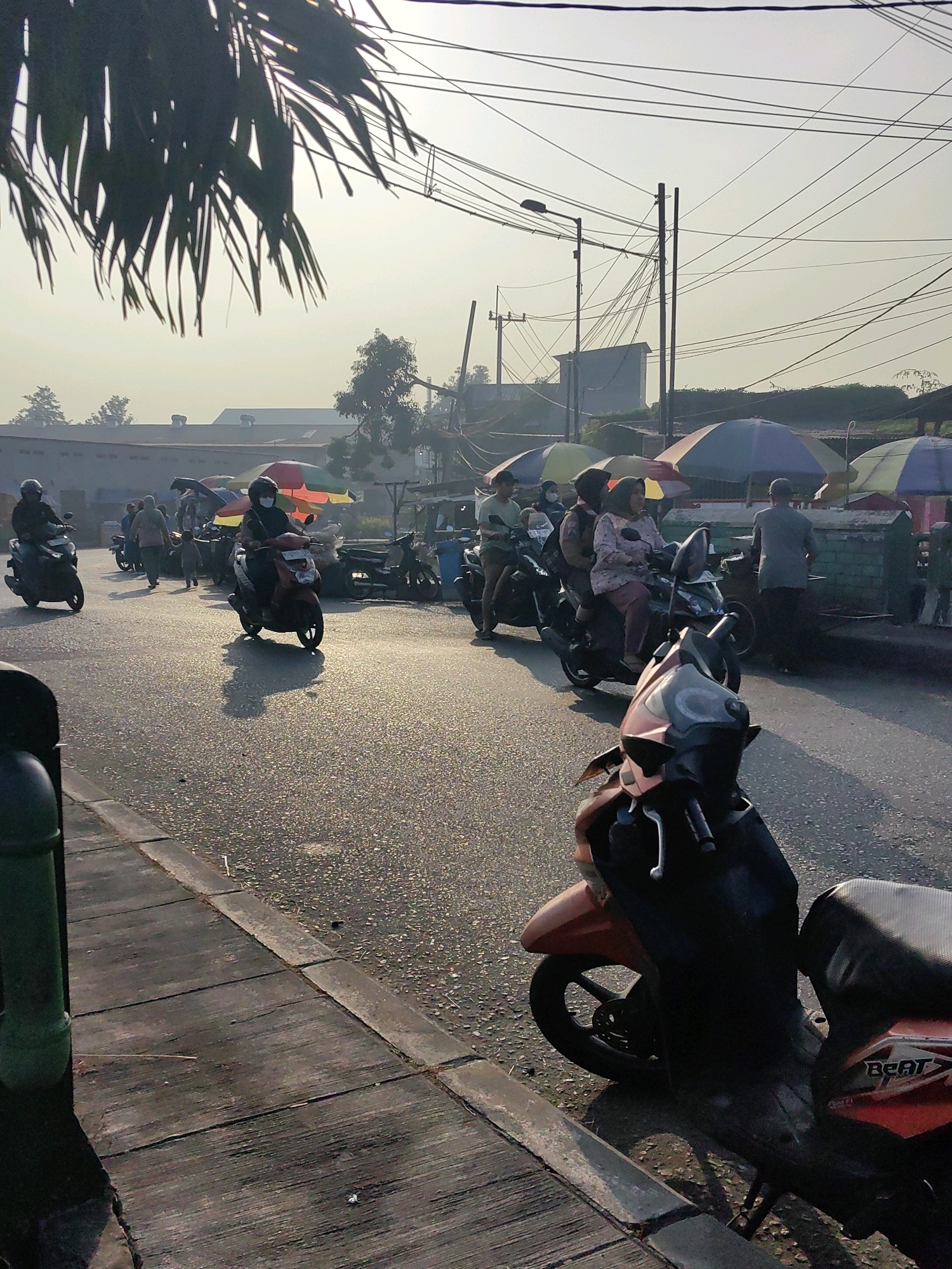 This is a photograph of a street scene in Keboansikep, Indonesia, taken during the day, possibly in the early morning or late afternoon, indicated by the long shadows and the warm, hazy sunlight. The image captures a bustling street with several people on motorcycles, suggesting a typical mode of transportation in the area.
In the foreground, on the right side of the image, a red and black scooter is parked, facing towards the viewer. Its "BEAT" branding is visible on the rear panel. To the left of the scooter, a gray sidewalk made of wooden planks is visible, bordering the asphalt road. A green cylindrical bin is also present on the sidewalk.
The road itself is wet and reflective, with numerous small stones and debris scattered on its surface. The sunlight creates shimmering reflections on the wet asphalt, along with distinct shadows cast by the people and vehicles.
Several people are riding motorcycles, moving along the road. In the center of the image, a person wearing a white mask rides a red scooter, with another person seated behind them. To their left, another scooter with a rider is visible, heading in the same direction. Further down the road, more motorcycles and people are present, along with several colorful umbrellas, some of which are open and provide shade for market stalls or vendors.
In the background, buildings with light-colored facades are visible, along with trees and dense vegetation. A complex network of overhead power lines and poles crisscrosses the sky, a common sight in urban and semi-urban environments in Indonesia. The sky is bright and hazy, with a pale yellow or white hue, suggesting the presence of dust or a light smog.
The overall impression is one of an active street in a community, with daily life unfolding amidst the infrastructure and natural elements of Keboansikep.