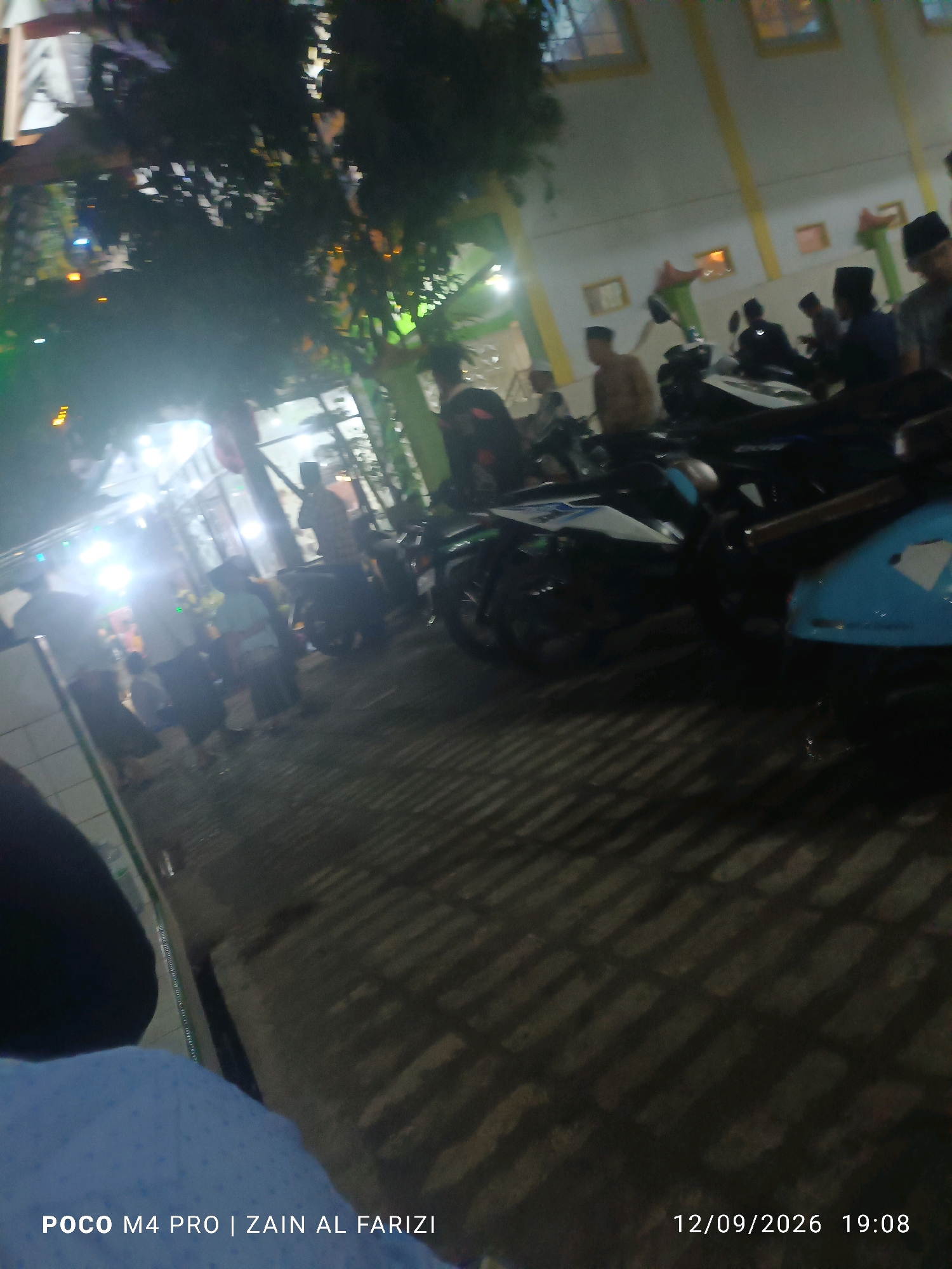 This is a photograph taken at night, presumably at a public gathering or event, possibly a religious one, given the presence of individuals wearing peci (caps often worn by Muslim men) and the dimly lit, outdoor setting with some artificial lighting. The image shows several motorcycles parked in a row, indicating transportation. A building with yellow trim and multiple windows is visible in the background. The ground is paved and shows distinct shadows cast by overhead structures and possibly trees. There are numerous people in the frame, mostly blurred due to motion or low light, but some appear to be standing or walking around the parked motorcycles. The overall atmosphere is one of a community event taking place after dark. The timestamp in the lower right corner indicates the photo was taken on September 12, 2026, at 19:08. The text in the lower left corner, "POCO M4 PRO | ZAIN AL FARIZI," suggests the camera model and possibly the photographer's name. The specific location in the city of Cendagah, Indonesia, cannot be determined from the visual information alone, but the presence of the motorcycles and the attire of some individuals suggest a locale where such cultural practices are common.
