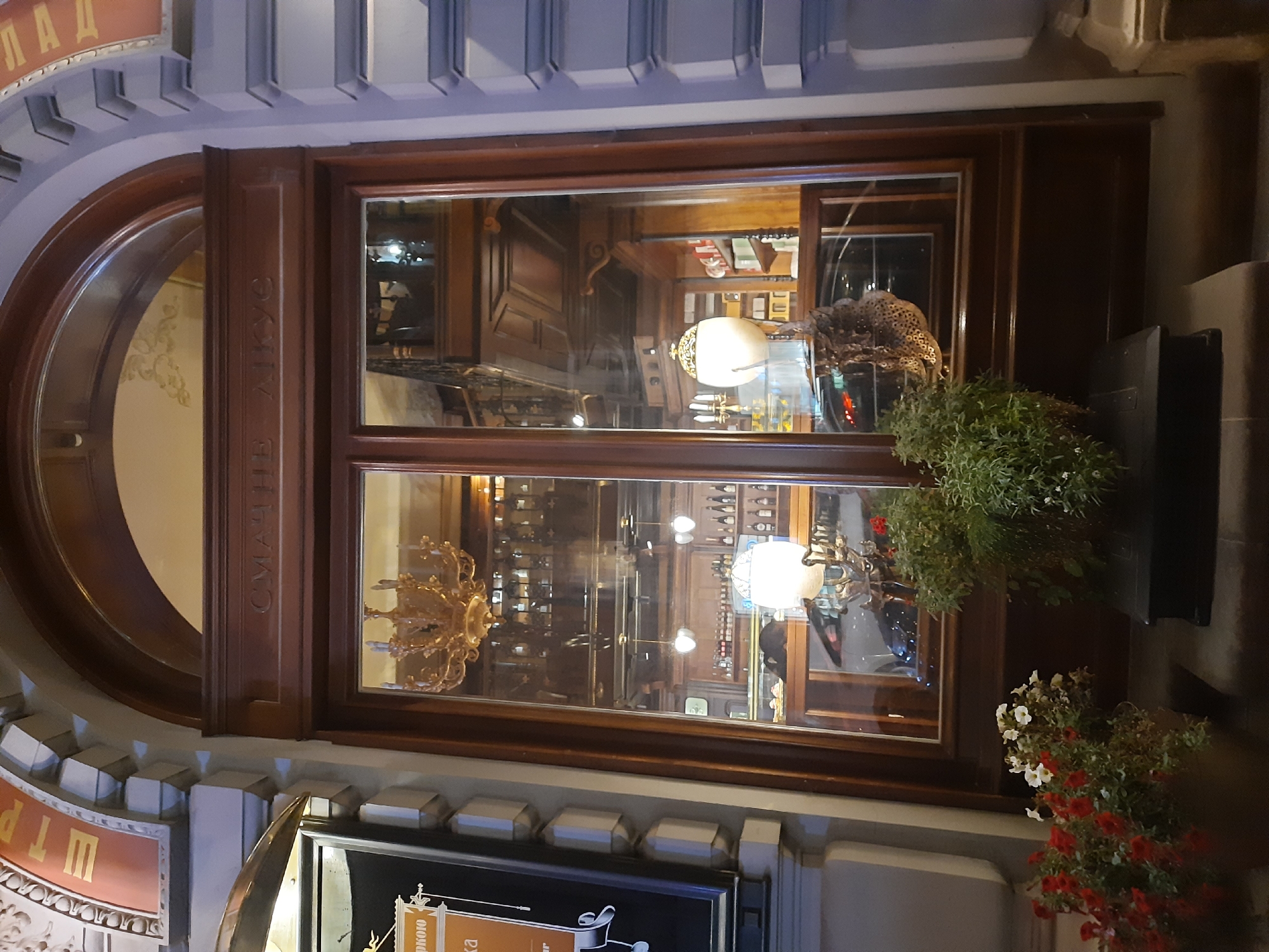 The image shows the storefront of "Smachno Lykous", a restaurant in Lviv, Ukraine. The storefront features a large, arched wooden door with two glass panes. Inside, a chandelier hangs from the ceiling, and shelves are stocked with bottles. In the foreground, two potted plants with red and white flowers are visible. The building's exterior is decorated with ornate carvings.