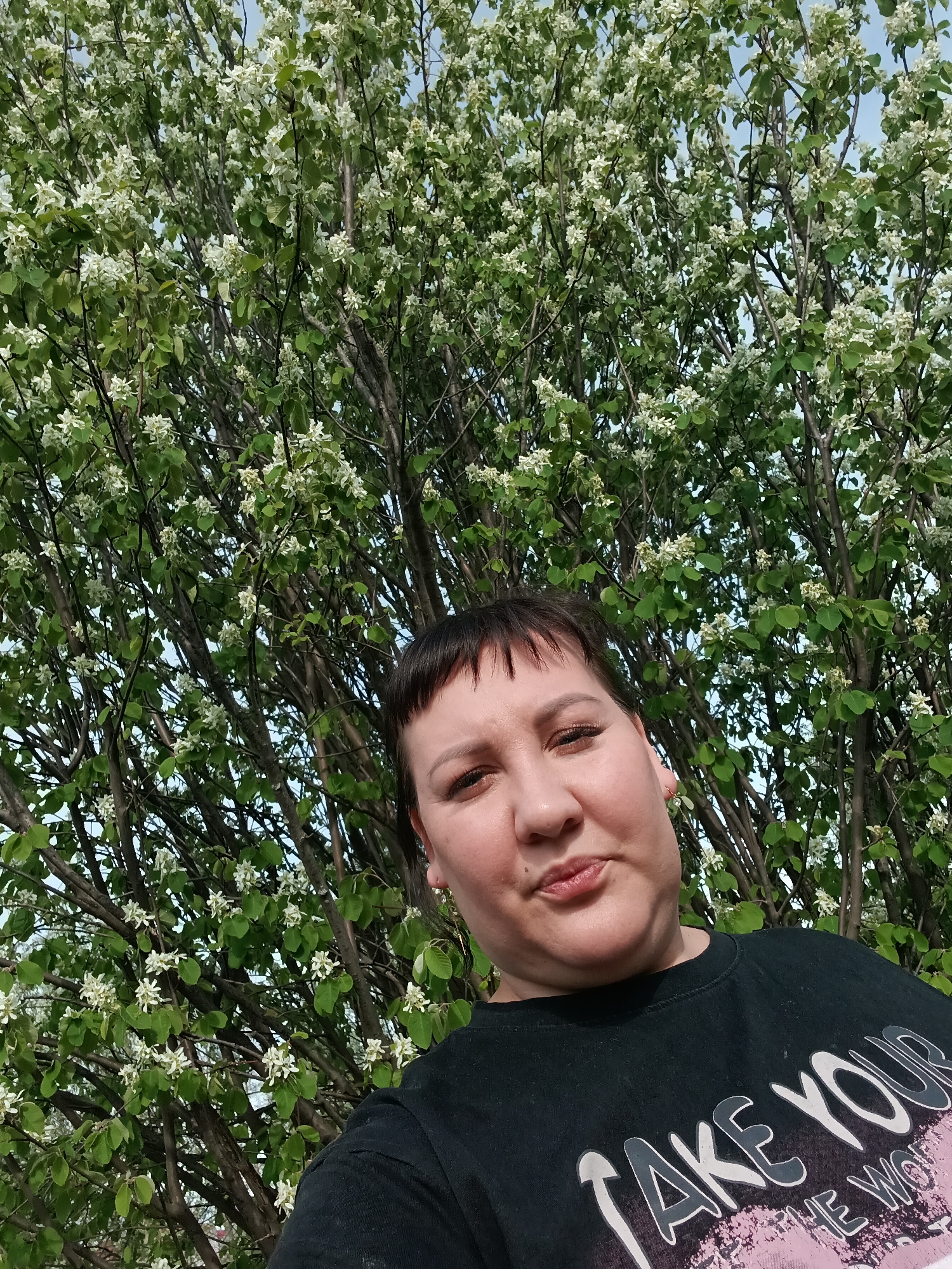 A woman stands in front of a flowering tree in Osa, Russia. The tree is covered in white blossoms and green leaves. She is wearing a black t-shirt with the words "Take Your The World" printed on it. The sky is visible through the branches of the tree.