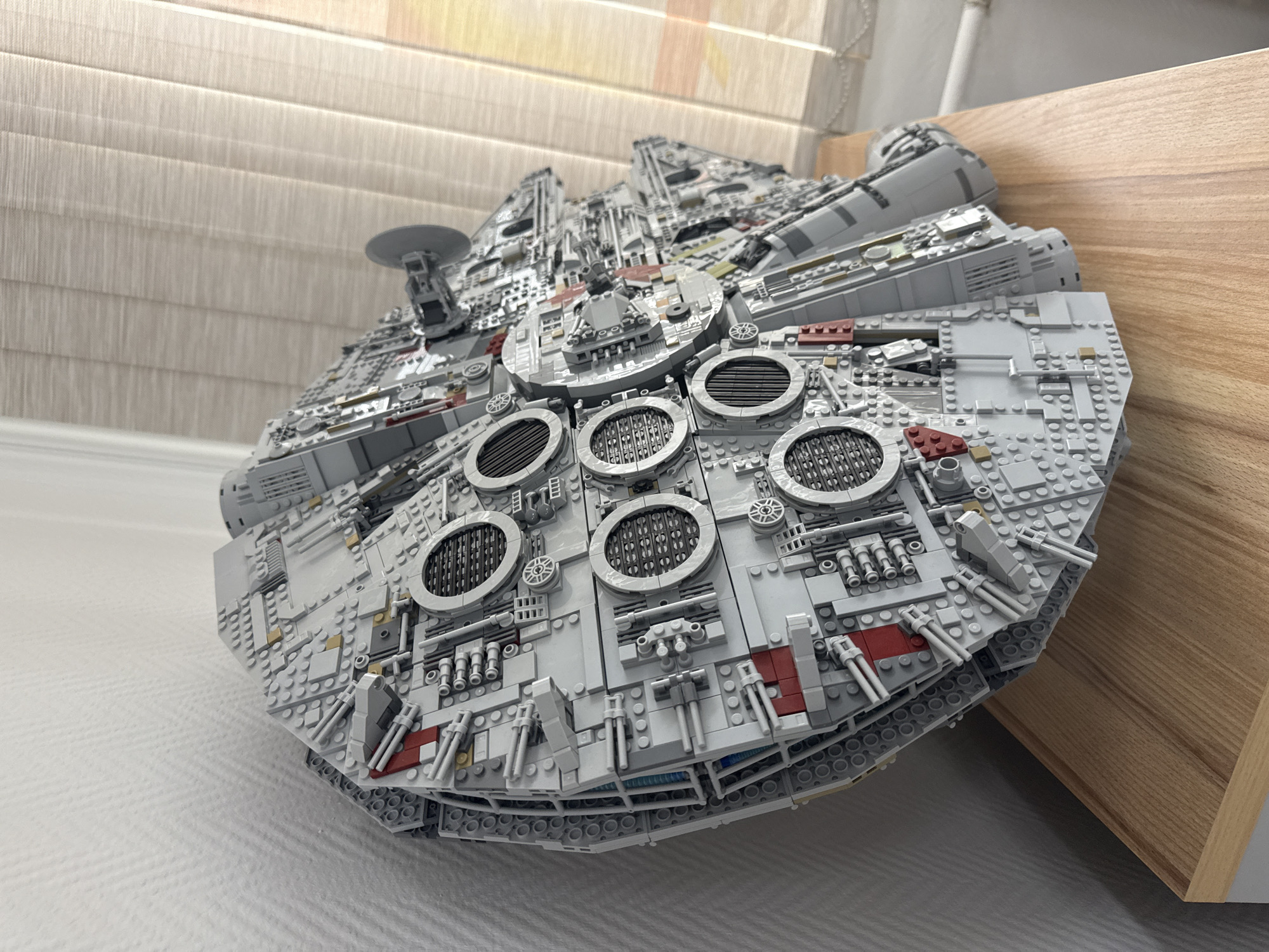 A LEGO Millennium Falcon model is the focal point of the image. The model is predominantly gray, with intricate detailing, including circular vents, guns, and a radar dish. It rests on a gray and white surface, possibly a floor, and is partially propped against a light-colored wooden structure. The background features neutral-toned vertical blinds. The location is not identifiable as Krasnoyarsk, Russia.