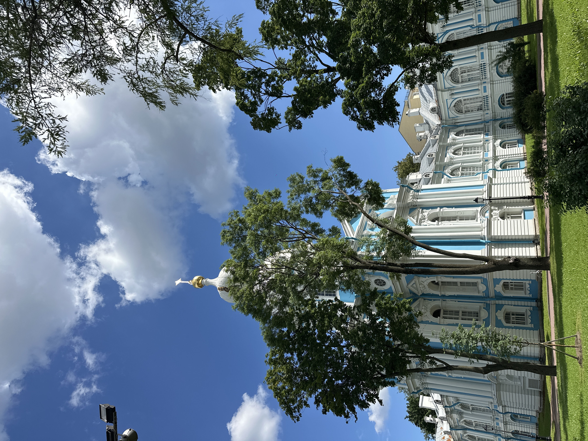 This image captures a sunny day in Saint Petersburg, Russia, featuring the Smolny Cathedral. The cathedral, a prominent landmark, is adorned in white and sky-blue with ornate white detailing. Its dome, topped with a golden cross, stands out against a vibrant blue sky dotted with fluffy white clouds. Lush green trees partially obscure the view of the cathedral, their leaves creating a dappled effect on the building and the manicured lawn in the foreground. A lamppost is visible on the left side of the frame, and a security camera is mounted on a pole near the top left corner. The architectural style is Baroque, characterized by its elaborate ornamentation and grandeur. The scene evokes a sense of peace and historical significance.