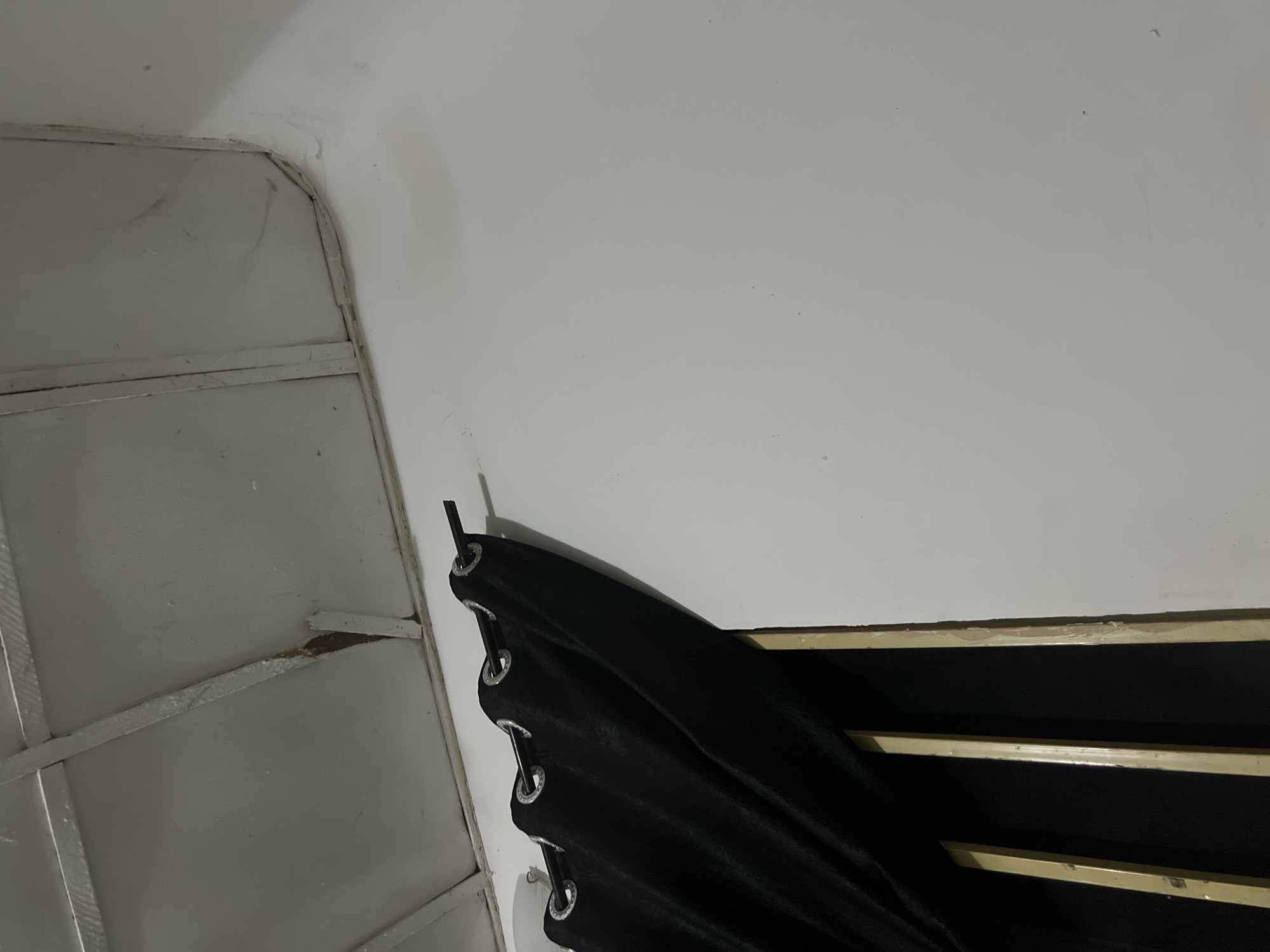 The image depicts a close-up view of a white wall and a section of a ceiling in what appears to be an indoor setting. The ceiling is constructed with white panels and exposed supporting beams, showing signs of wear and tear with some damage and peeling paint visible. A dark curtain with silver grommets is partially visible, hanging from a dark rod. The curtain covers a portion of a window or opening, through which some vertical bars are visible. The wall is a plain white surface, with some minor marks or scuffs. The scene does not contain any people or indicate any specific actions or interactions. The location is specified as Howayi, Nigeria.