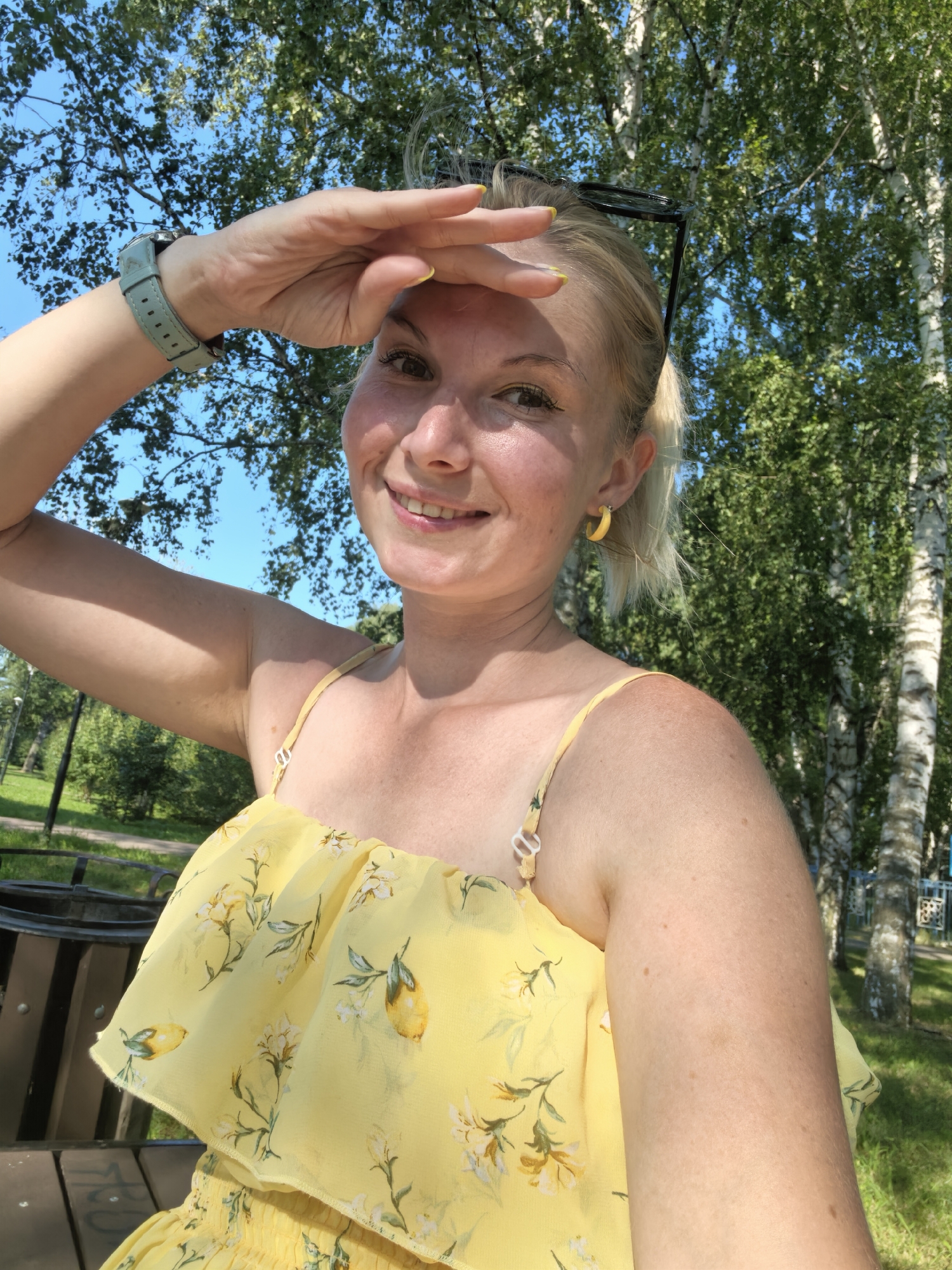 A blonde woman is taking a selfie outdoors in a park. She is wearing a yellow dress with a lemon print and yellow hoop earrings. She is shielding her eyes from the sun with her hand. In the background, there are trees, a blue sky, and a trash can. The image appears to have been taken in the city of Sterlitamak, Russia.