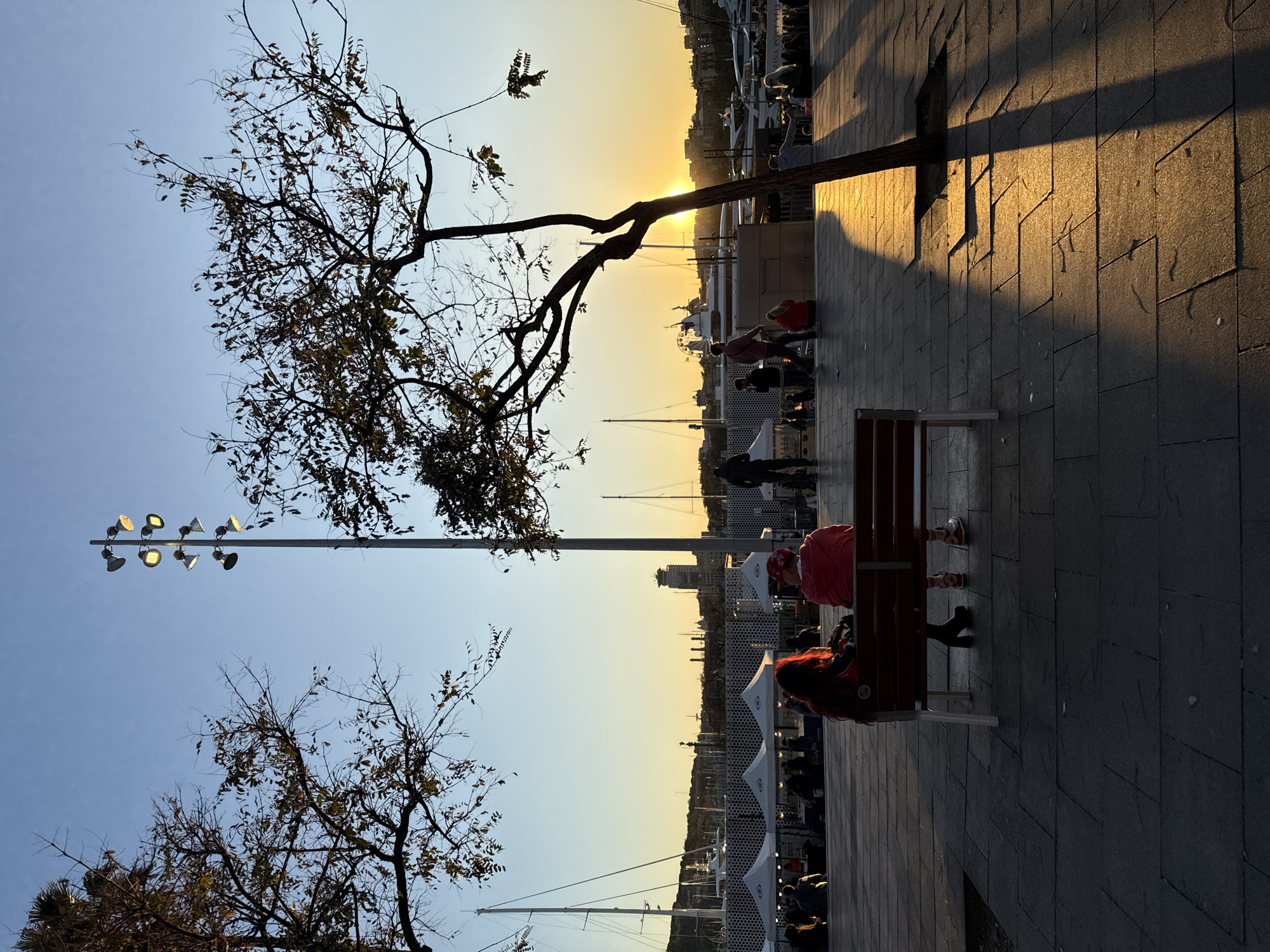 The image shows a waterfront scene at sunset in Barcelona, Spain. Silhouetted figures of people sit on benches along a paved walkway. Sailboats and other watercraft are visible in the harbor. A bare tree and a light post with multiple lamps are prominent on the left. The sun sets behind the harbor, casting long shadows on the pavement.