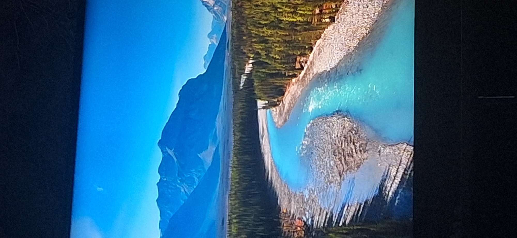 The image depicts a wide, turquoise river winding through a dense evergreen forest. In the background, majestic, snow-capped mountains rise against a clear blue sky. The riverbanks are lined with pebbles and rocks, and on the left side, a sandy area shows distinct ripples from the water's flow. There is a small building nestled amongst the trees on the right bank. The overall scene is a serene and picturesque natural landscape. There is no indication of the city of Bila Tserkva, Ukraine in this image.