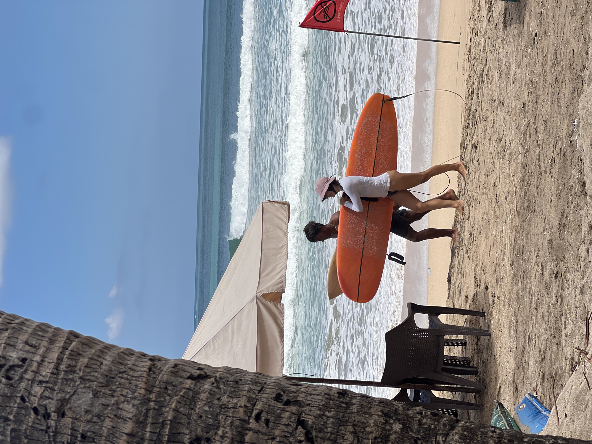 The image depicts a beach scene in Kuta, Indonesia, with two individuals carrying surfboards near the ocean. A large palm tree trunk partially obscures the left side of the frame, with its textured bark prominently visible. Behind the tree, a beige beach umbrella provides shade for two dark plastic chairs. The sand in the foreground is a light brown color, with scattered debris.
The ocean is a vibrant turquoise blue with moderate waves breaking on the shore. The sky above is clear and bright blue with a few wispy white clouds. To the right of the image, a red flag with a black symbol and text reads "SWIMMING FORBIDDEN," indicating a potential hazard or restriction in the water.
The two individuals are walking on the sand, carrying an orange surfboard between them. The person on the left appears to be a male, wearing dark shorts and no shirt. The person on the right is wearing a white long-sleeved rash guard, black bikini bottoms, and a light pink hat. They are holding the surfboard by its sides. Another lighter-colored surfboard is positioned behind the orange one, resting against the legs of the individuals. The overall scene suggests a typical day at a popular Indonesian beach.
