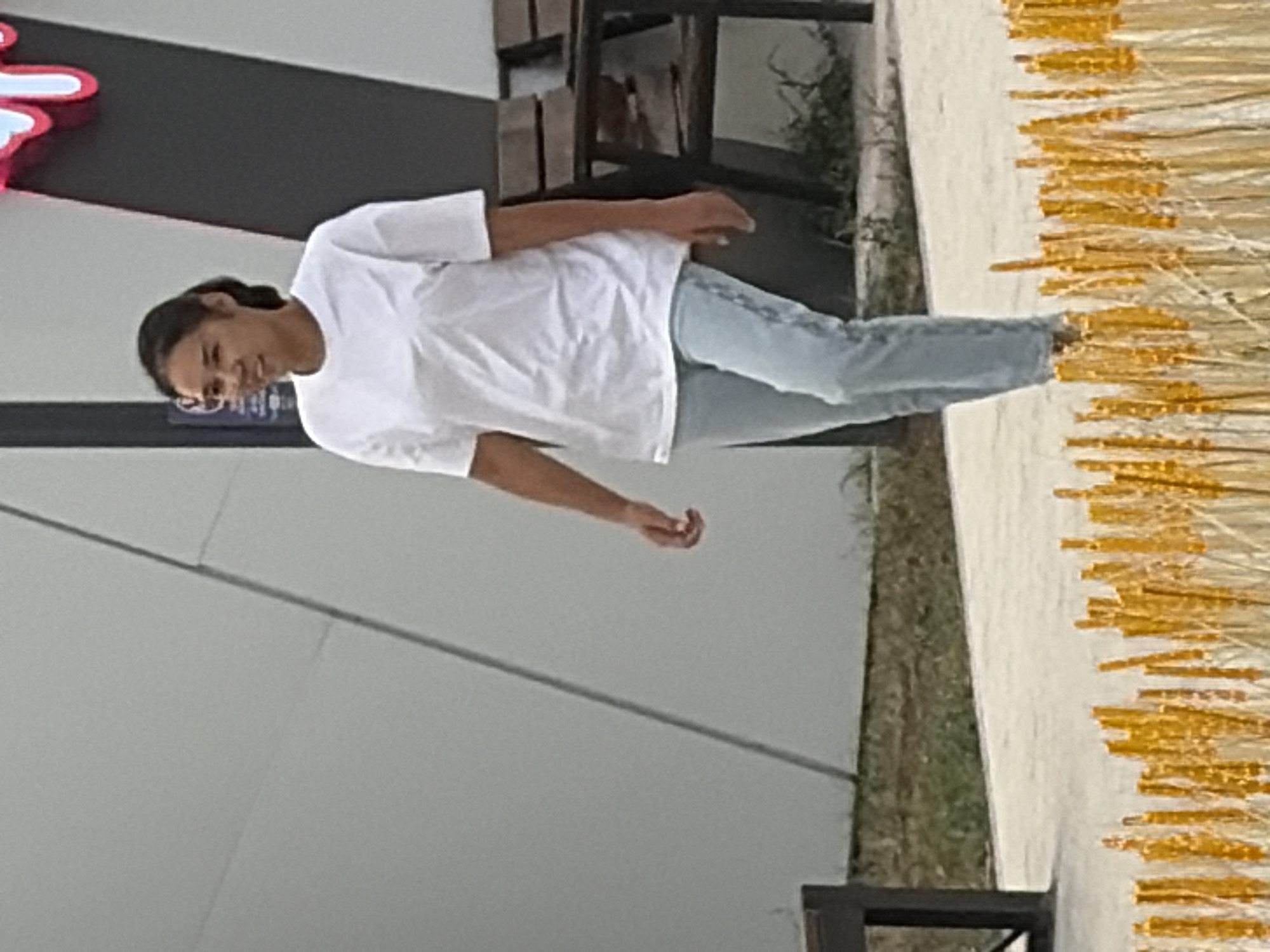 A medium shot captures a young woman with dark hair pulled back into a ponytail, walking towards the right of the frame, her head turned to her left as if looking at something off-camera. She is wearing a plain white t-shirt and light-wash denim jeans. The background consists of a light gray, angular building with a dark vertical element to its right, featuring a bright, stylized pink and white graphic. In the middle ground, a wooden bench with dark metal legs is visible. The foreground is partially obscured by a decorative arrangement of golden-yellow, stick-like objects. The ground is a pale gray pathway. The lighting suggests an outdoor setting during the daytime, with soft shadows.