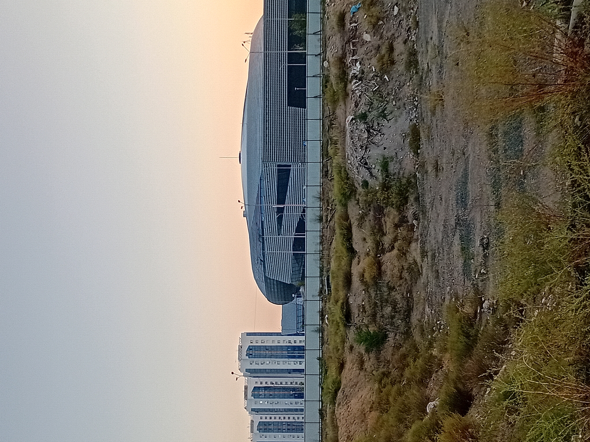 The image depicts the Sary-Arka Velodrome in Semey, Kazakhstan. In the foreground, a patch of dry, overgrown vegetation and dirt covers uneven terrain. A dark metal fence runs horizontally across the mid-ground, separating the foreground from a paved area behind it. Beyond the fence, a long, modern building with a curved, metallic facade is visible. This building appears to be a sports facility or arena, characterized by its sleek design and horizontal paneling. To the left of this main structure, three multi-story residential buildings stand in a row, featuring a light-colored facade with blue and darker accents. The sky above is a soft gradient of pale blue and orange, suggesting it is either dawn or dusk. No people are visible in the frame.