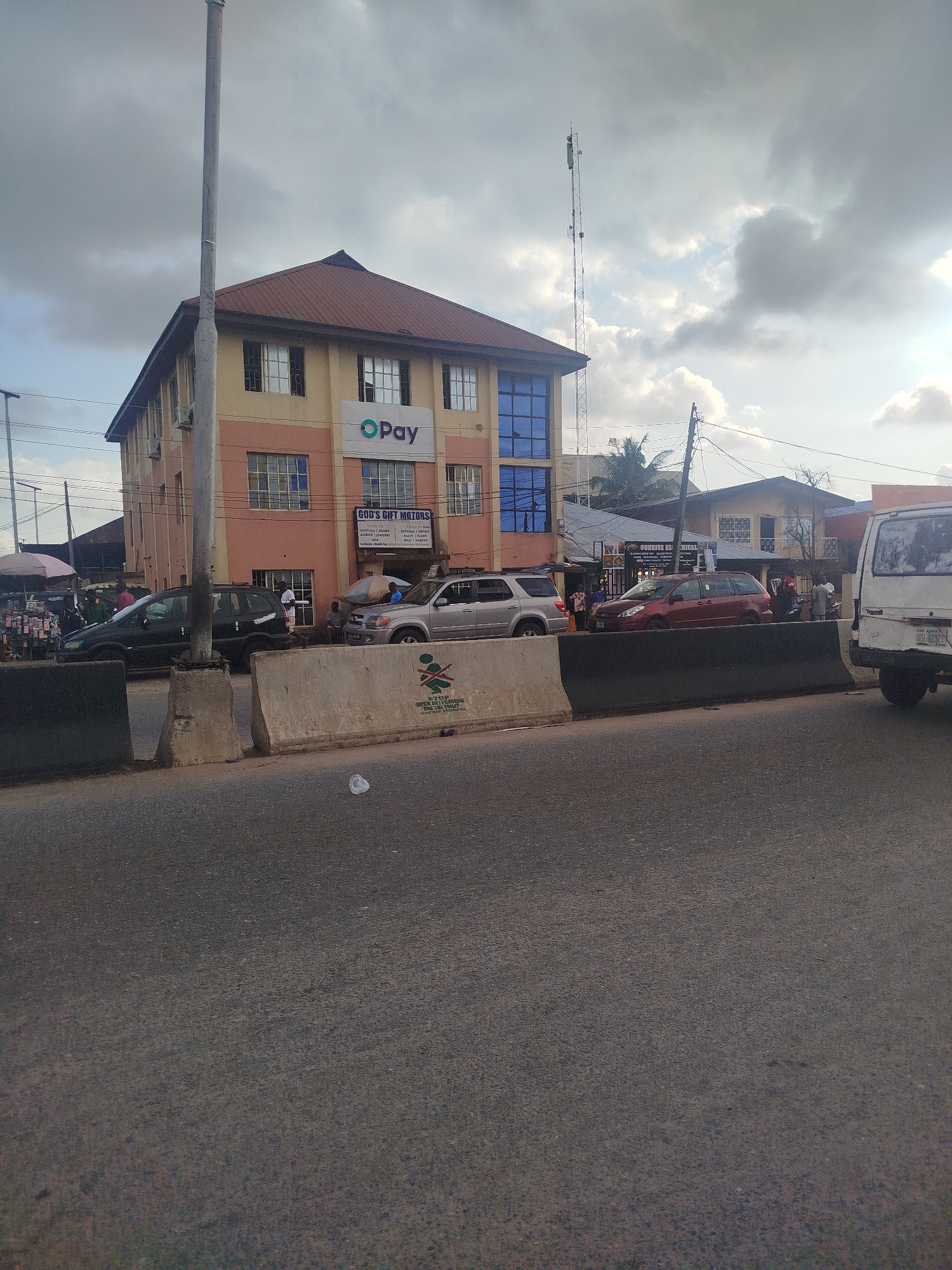 The image depicts a street scene in Benin City, Nigeria, focusing on a multi-story building with a prominent "O'Pay" logo and another sign for "GOD'S GIFT MOTORS." The building has a beige and pink facade with multiple windows, some of which are tinted blue. A large cell tower is visible behind the building, reaching high into the overcast sky. In front of the building, on the street, are several vehicles, including a black SUV, a silver SUV, a red minivan, and a white van. People are seen standing around the vehicles and on the sidewalk. Concrete barriers divide the roadway, and a street vendor with an umbrella is present to the left. The overall environment appears to be a busy urban commercial area.