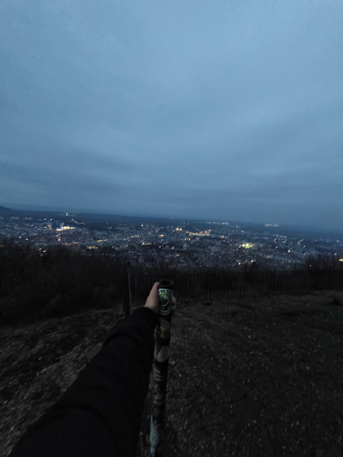 The image captures an elevated panoramic view of the city of Shumen, Bulgaria, during dusk or night, under a dark blue-gray, overcast sky. In the immediate foreground, a human arm, clad in a dark long-sleeved garment, extends from the lower-left, holding a cylindrical object vertically. The object displays a green label with white text including "SK 200" and an indistinct emblem, with a camouflage-like pattern on its lower body. The ground beneath the arm is dark and uneven, consistent with sparse earth or low vegetation on a hillside. A dark, low wooden fence runs horizontally across the mid-ground, separating the vantage point from the city below. The background features a sprawling cityscape illuminated by numerous scattered artificial lights, appearing as bright pinpoints against the dark urban mass. Distant hills or landforms are vaguely discernible at the horizon, below the extensive cloud cover.