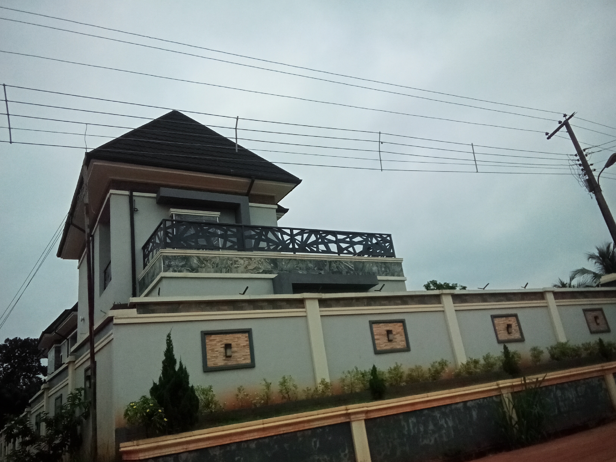 The image depicts a modern two-story house with a grey facade and a dark-tiled pyramidal roof, located in Oko Town, Nigeria. The house is partially visible on the left side of the frame, with a tall, narrow structure featuring multiple windows and air conditioning units. A prominent grey concrete wall with decorative square insets, each containing textured brick-like material, surrounds the property. A black metal railing with a geometric pattern adorns the balcony of the upper level of the house, visible above the wall. The sky is overcast and grey. Electrical wires are strung across the upper portion of the image, supported by utility poles, with one pole clearly visible on the right side. Small trees and bushes are planted along the interior side of the wall. The foreground shows a reddish-brown surface, possibly dirt or a pathway.