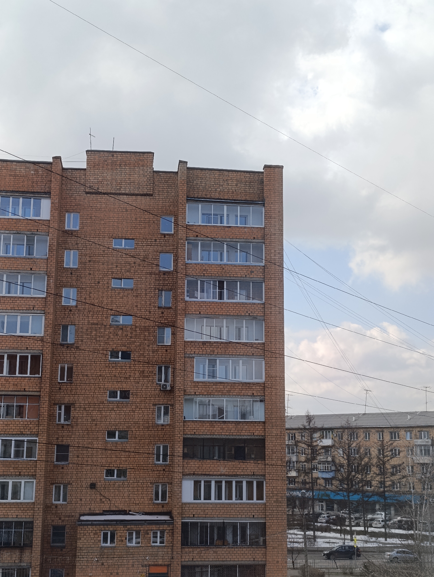 The image shows a brick apartment building in Krasnoyarsk, Russia. The building is several stories high and has many windows, some with balconies. There are wires running across the sky in front of the building. Another similar building is visible in the background, partially obscured by leafless trees. Cars are parked along the road in front of the building. The sky is cloudy.