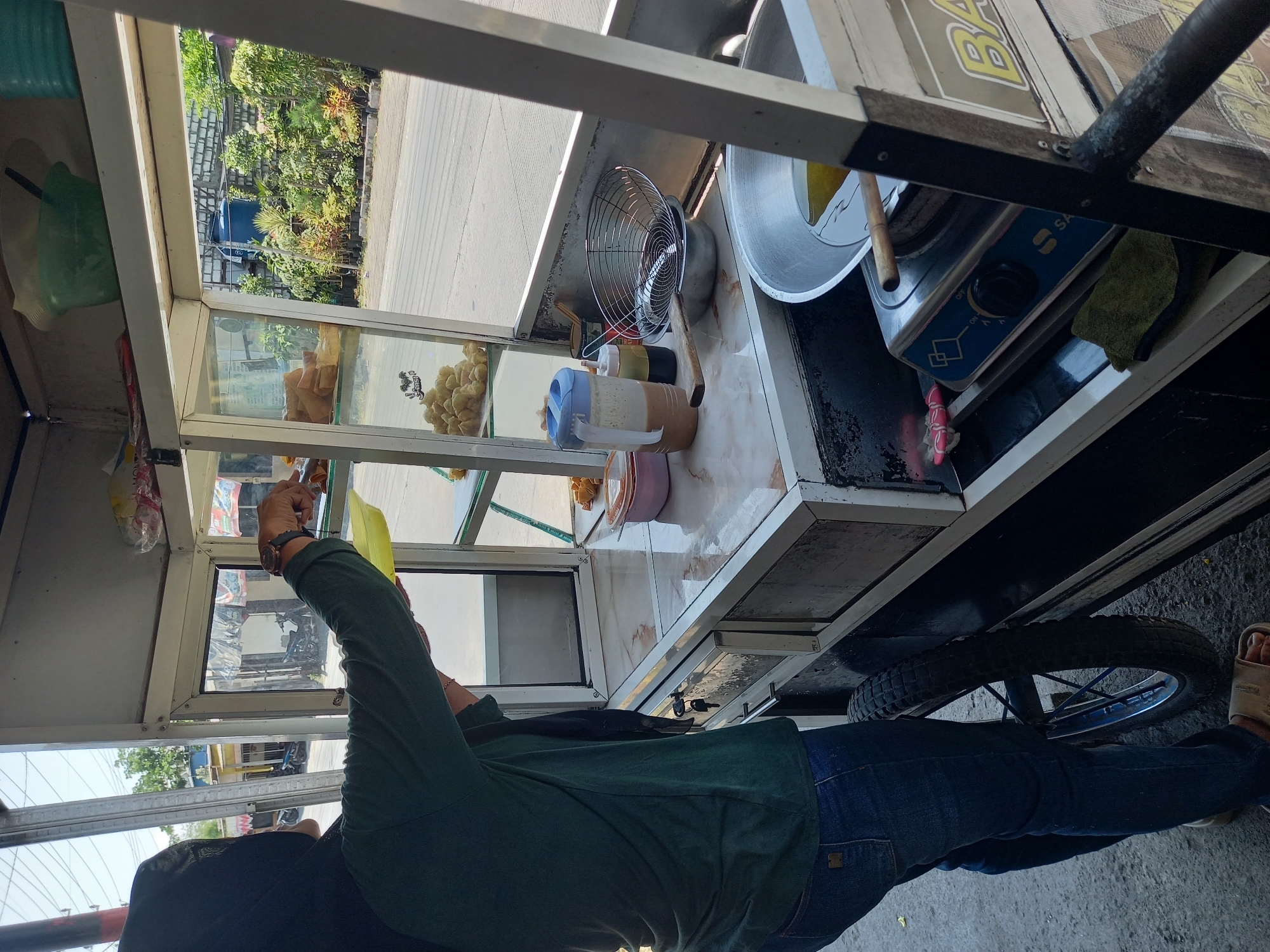 In the city of Pelemwatu, Indonesia, a person wearing a dark green long-sleeved shirt, blue jeans, and a black hijab stands beside a food cart. The person is holding a yellow plastic tray in their right hand and appears to be interacting with the contents of the food cart's display case. The food cart has a glass display with shelves containing food items and a lower section with drawers and cooking equipment. On the counter of the cart, there are containers, a pitcher, a strainer, and a wok with oil on a portable stove. The background shows a street scene with buildings, trees, and other vehicles. The overall environment suggests a street food vendor operation in a sunny outdoor setting.