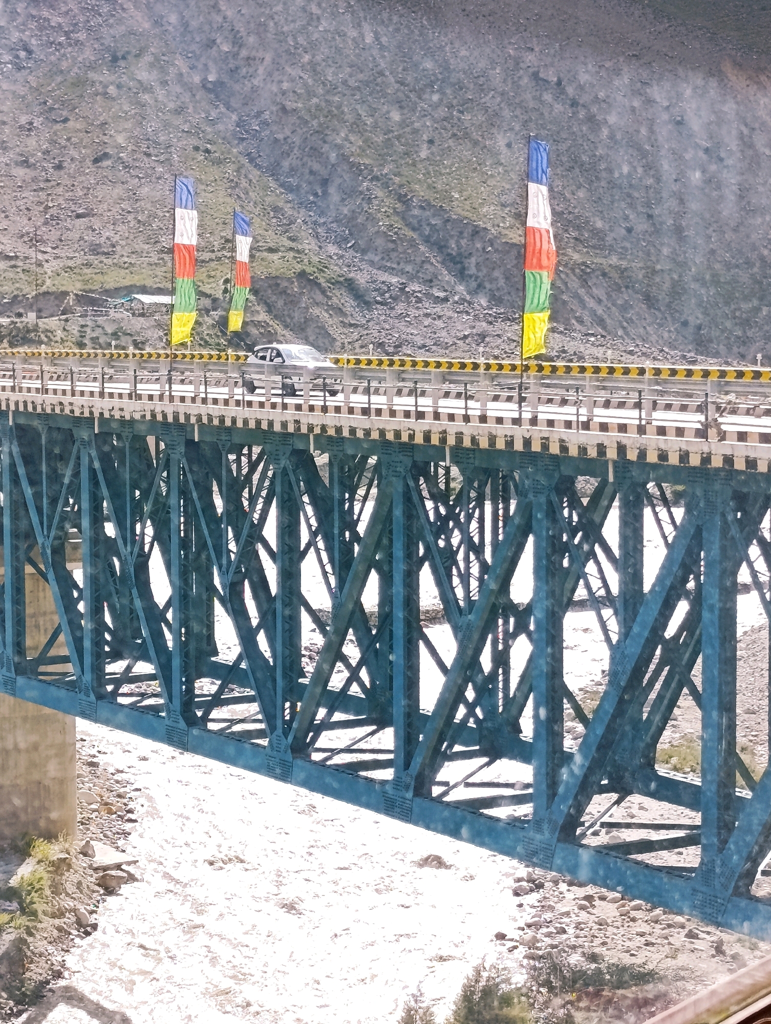 The image captures a wide, shallow river flowing beneath a substantial blue metal truss bridge. The riverbed is rocky, with the water appearing murky and light brown, suggesting a high sediment content. The bridge itself is a prominent structure with a latticework of steel beams. The bridge deck is paved and has railings. On the bridge, a silver car is visible, driving from left to right. Along the side of the bridge, colorful prayer flags are strung vertically, adding a cultural element to the scene. The flags are red, white, blue, green, and yellow. In the background, steep, rocky hills rise up, sparsely vegetated. The overall environment appears to be a mountainous region. The image was taken on June 27, 2024, in Darcha, India.