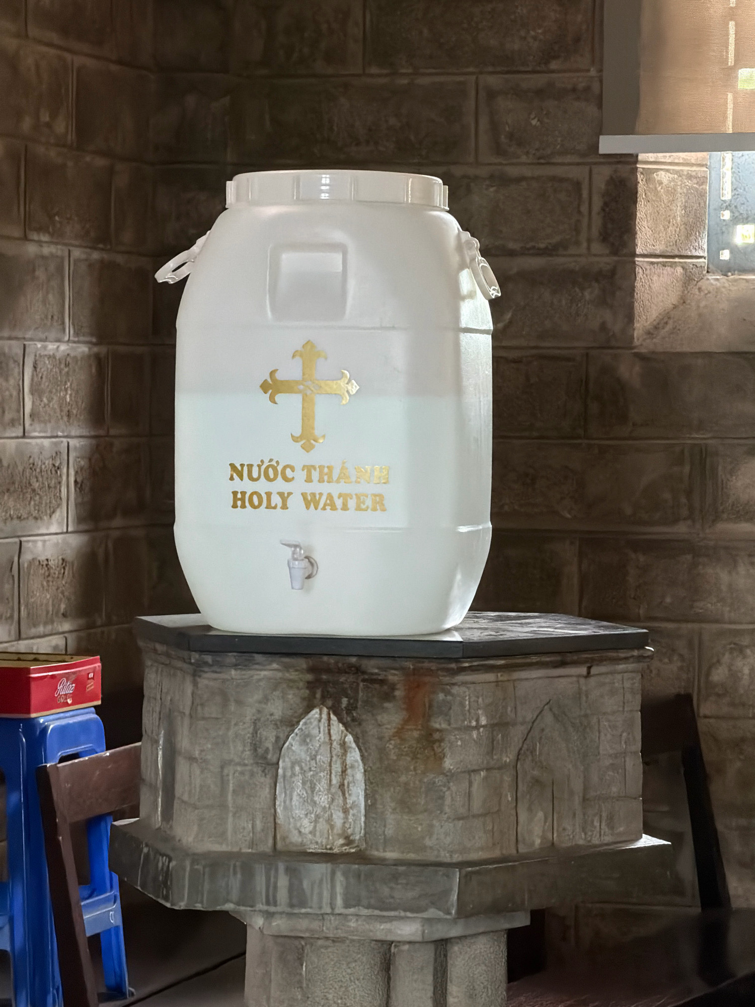 A large, translucent white plastic container filled with clear liquid sits atop a stone pedestal. A golden cross and the text "NƯỚC THÁNH HOLY WATER" are affixed to the front of the container. A small spigot protrudes from the lower front of the container. The pedestal has a decorative arched indentation on its front. Behind the container and pedestal, a stone brick wall forms the background. To the left of the pedestal, a blue plastic stool and a wooden chair are partially visible, with a red box on top of the stool. A window with diffused light is visible in the upper right corner. The scene appears to be an interior setting within a church or religious building. This scene is located in Nha Trang, Vietnam.