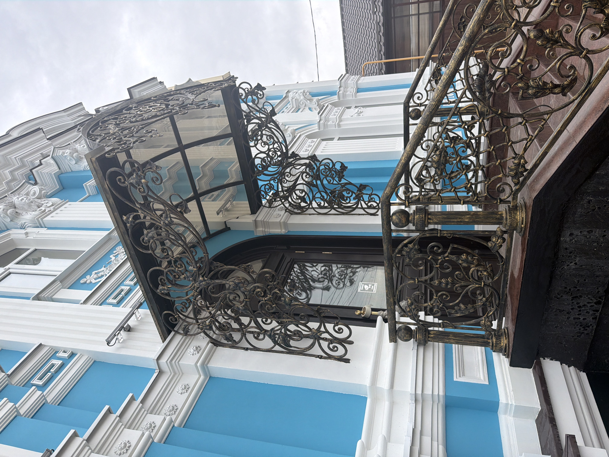 The image shows a section of a building located in Leningradskaya, Russia. The building is painted in a distinctive color scheme of blue and white, with detailed architectural moldings and decorative elements. A wrought-iron canopy with intricate scrollwork extends over a door or window. A staircase with ornate iron railings runs alongside the building. The sky is overcast.