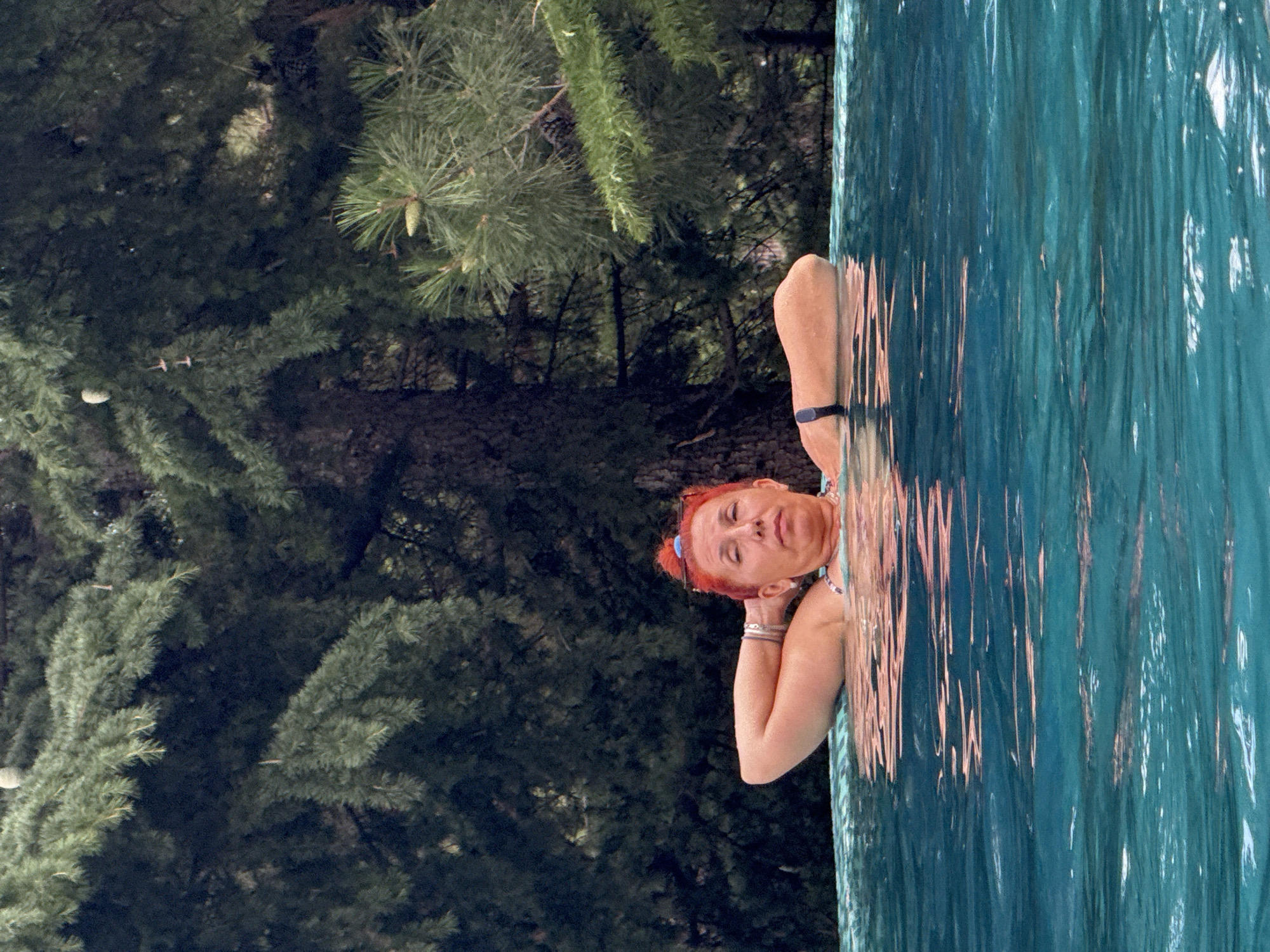 A woman with red hair is pictured from the chest up, submerged in turquoise water, resting her arms behind her head. She appears to be in a swimming pool, possibly an infinity pool, as the water surface extends to the foreground with reflections of the sky and surrounding trees. The background is dominated by dense, dark green coniferous trees. The woman is wearing a patterned swimsuit and a black fitness tracker on her left wrist. She has several bracelets on her right wrist. The lighting suggests an overcast or shaded environment. The image does not contain any identifiable landmarks or indicators of a specific city or location like Yalta, Ukraine.