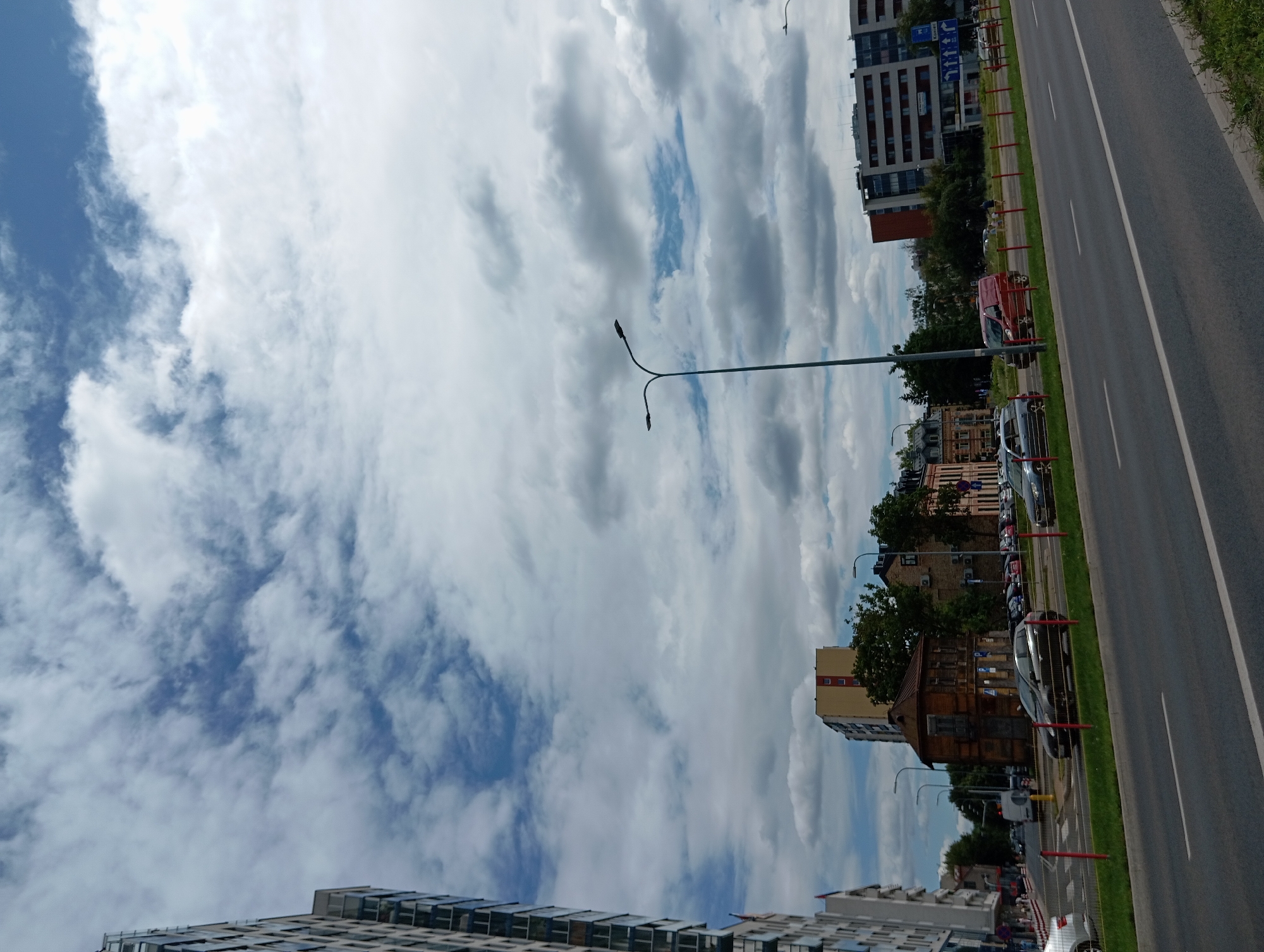 The image shows a street scene in the city of Białystok, Poland, under a cloudy sky. On the left, a tall, modern apartment building with many windows is visible. Across the street, several cars are parked along the side of the road. In the middle ground, there are older, low-rise buildings, including a brown, wooden structure and other brick buildings. Further down the street, a larger, modern building with a red facade is prominent. A street lamp with two lights stands in the foreground, and another one is visible in the distance. Cars are driving on the road, and some people can be seen walking on the sidewalk in the background. The road has multiple lanes and is bordered by a grass verge with red posts.