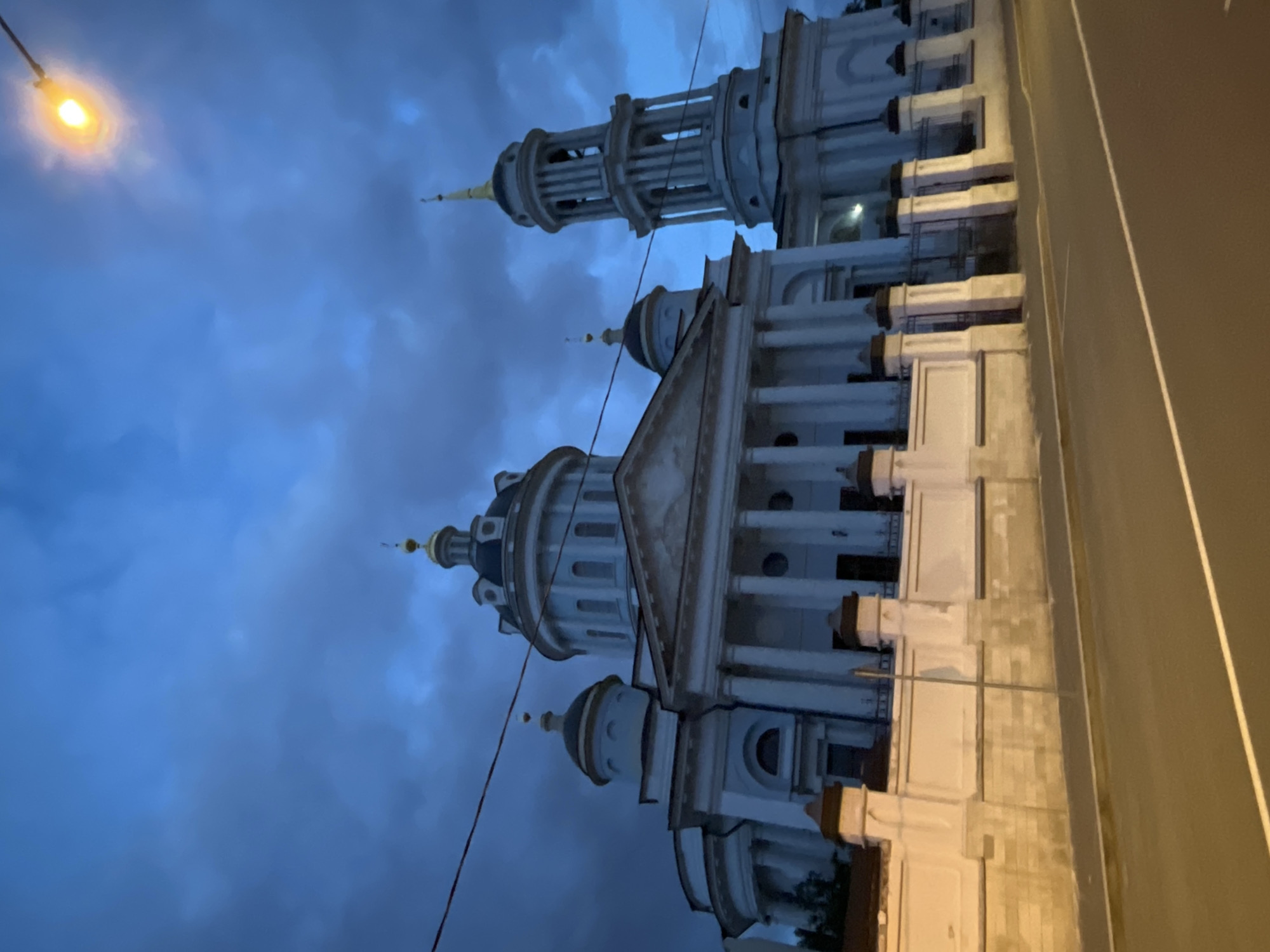 The image captures a white building with architectural details against a blue sky. The building features columns, domes, and a tower-like structure. A street lamp is visible in the upper left corner, and a street is adjacent to the building on the right side. The location is city of Unknown, Unknown.