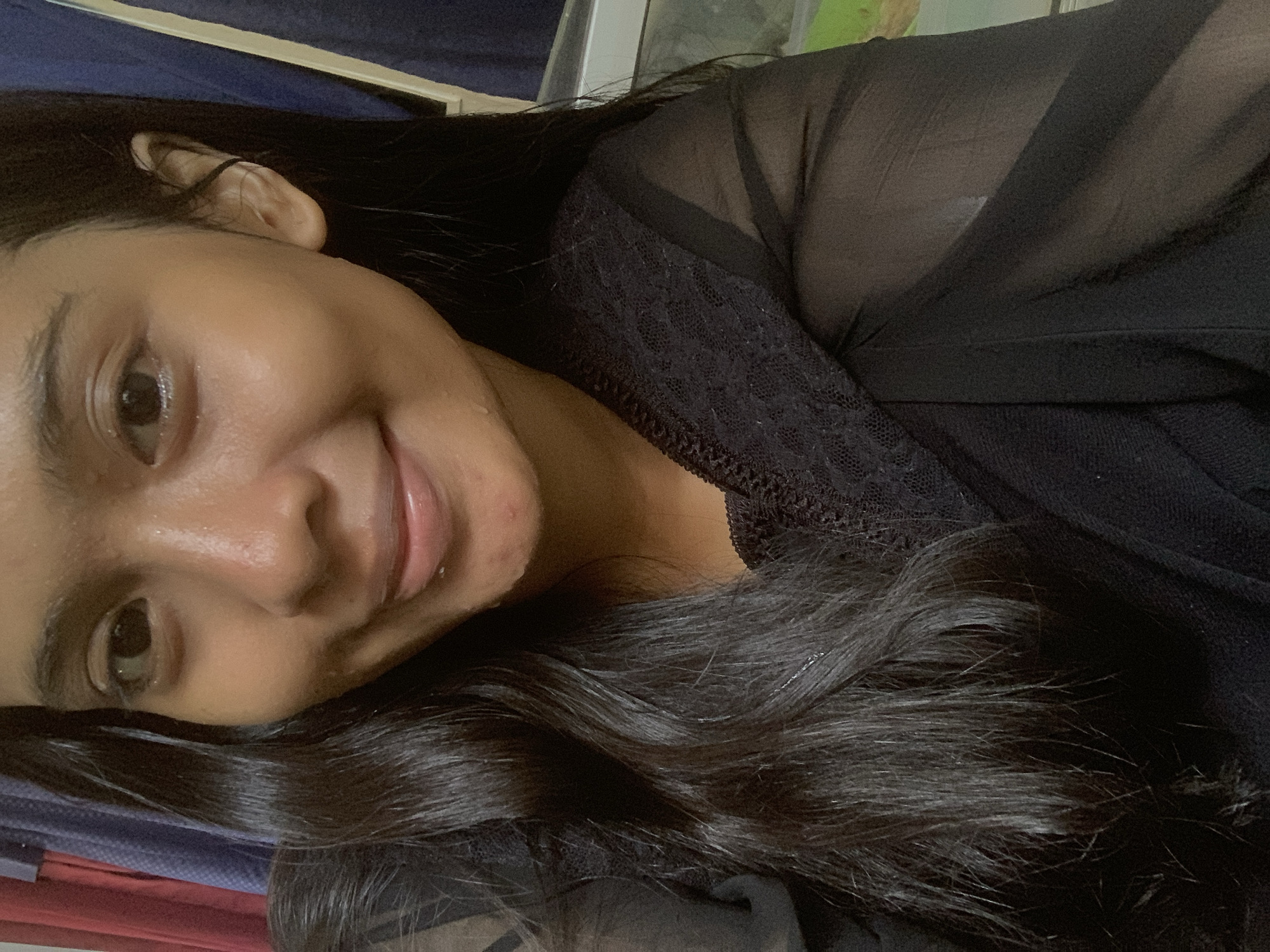 The image displays a close-up, tilted portrait of a young woman, likely in her late teens or early twenties, smiling gently while looking directly at the camera. Her head is angled, suggesting she is lying down, possibly taking a selfie. She has long, dark, wavy hair that frames her face and flows over her shoulder and chest. Her skin has a warm, tan complexion, with a noticeable sheen on her forehead and cheek. One dark eye is clearly visible, along with part of her nose and her smiling pink lips.
She is dressed in a black top. The upper section of the garment features a sheer, possibly lace or mesh fabric, which subtly reveals an underlying black garment, likely a bra or camisole. The lower part of her top is a solid black fabric.
The background is softly blurred, indicating an indoor setting. A dark blue fabric, possibly a bedsheet or cushion, is visible in the upper left portion of the image. Further back, some out-of-focus lighter shapes suggest furniture or elements of a room. The lighting is soft and even, typical of an indoor environment. No visible text is present in the image. This scene is located in Sungai Meriam, Indonesia.