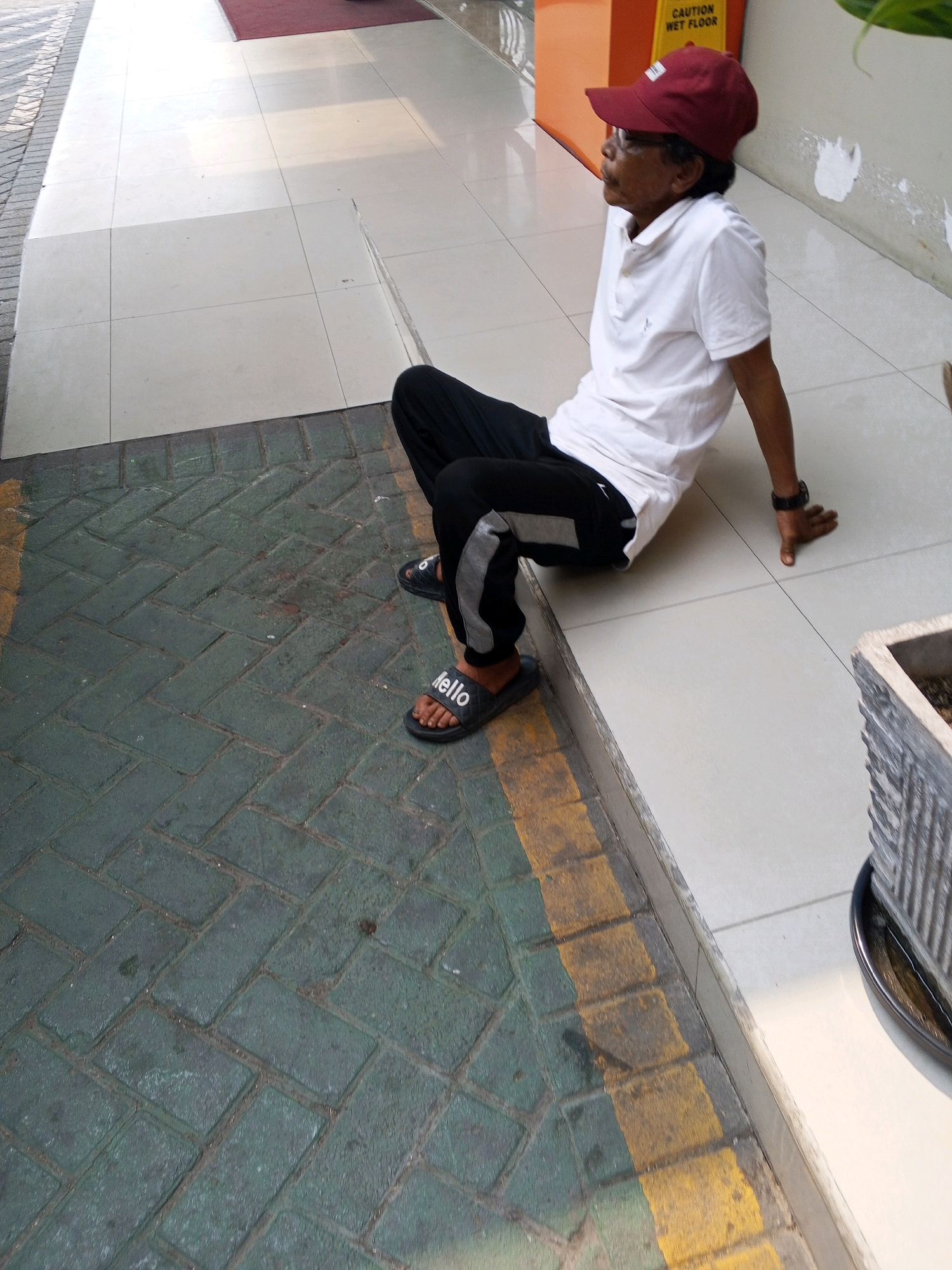 A man wearing a red cap, white polo shirt, and black pants with grey stripes is sitting on a tiled floor next to a caution wet floor sign. He is wearing black sandals with the word "hello" on them. The environment appears to be an outdoor walkway or public space. The flooring consists of light-colored tiles and dark green brick pavers with yellow painted lines. In the background, there is an orange caution sign and a partial view of a plant in a concrete pot. The lighting suggests it is daytime. The location is identified as Ciruas, Indonesia.