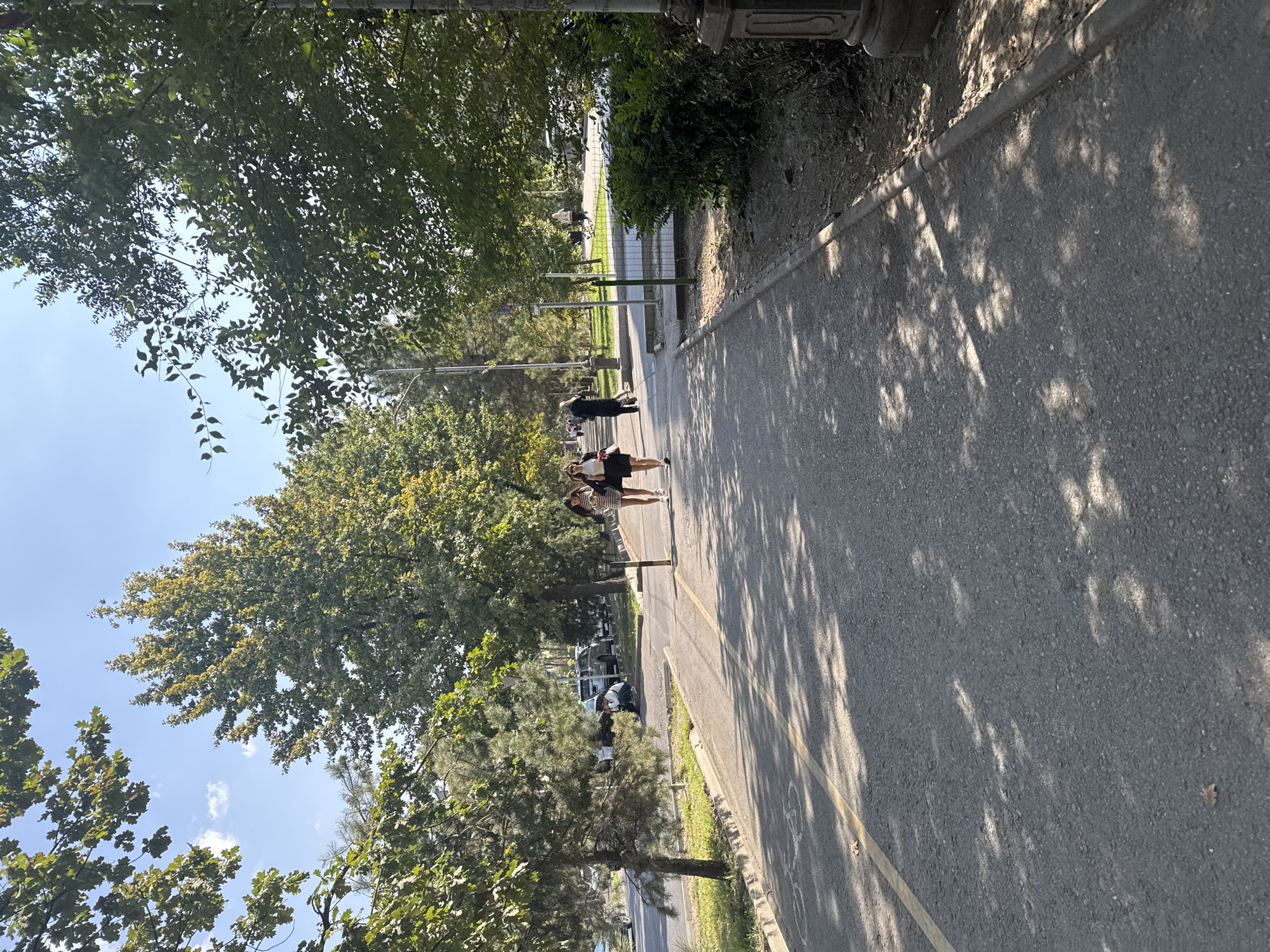 The image depicts a paved pathway lined with trees in Bishkek, Kyrgyzstan. Two young women are walking side-by-side on the pathway, dressed in casual attire. One woman is wearing a striped dress, and the other is in a black skirt and white top. They are walking towards the viewer's right. In the background, several cars are parked along the roadside, and other pedestrians are visible further down the path. The environment appears to be a park or a tree-lined street, with sunlight casting shadows on the pathway. The trees have green foliage, suggesting it is either spring or summer. The pathway has a yellow line marking its edge, and a bicycle symbol is visible on the asphalt.