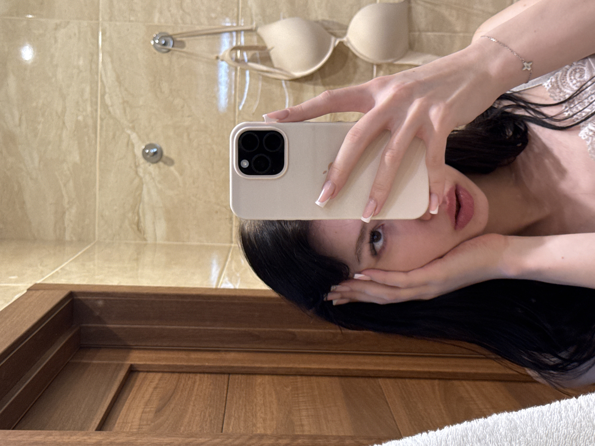 A young woman is taking a selfie in a bathroom, holding her phone in her right hand to capture her reflection. She has long, dark hair, and her left hand is resting on her face. She is wearing a light-colored top. In the background, there are beige tiled walls, a wooden door on the left, and a light-colored bra hanging on a hook on the right. The image appears to have been taken indoors, likely in a residential bathroom.